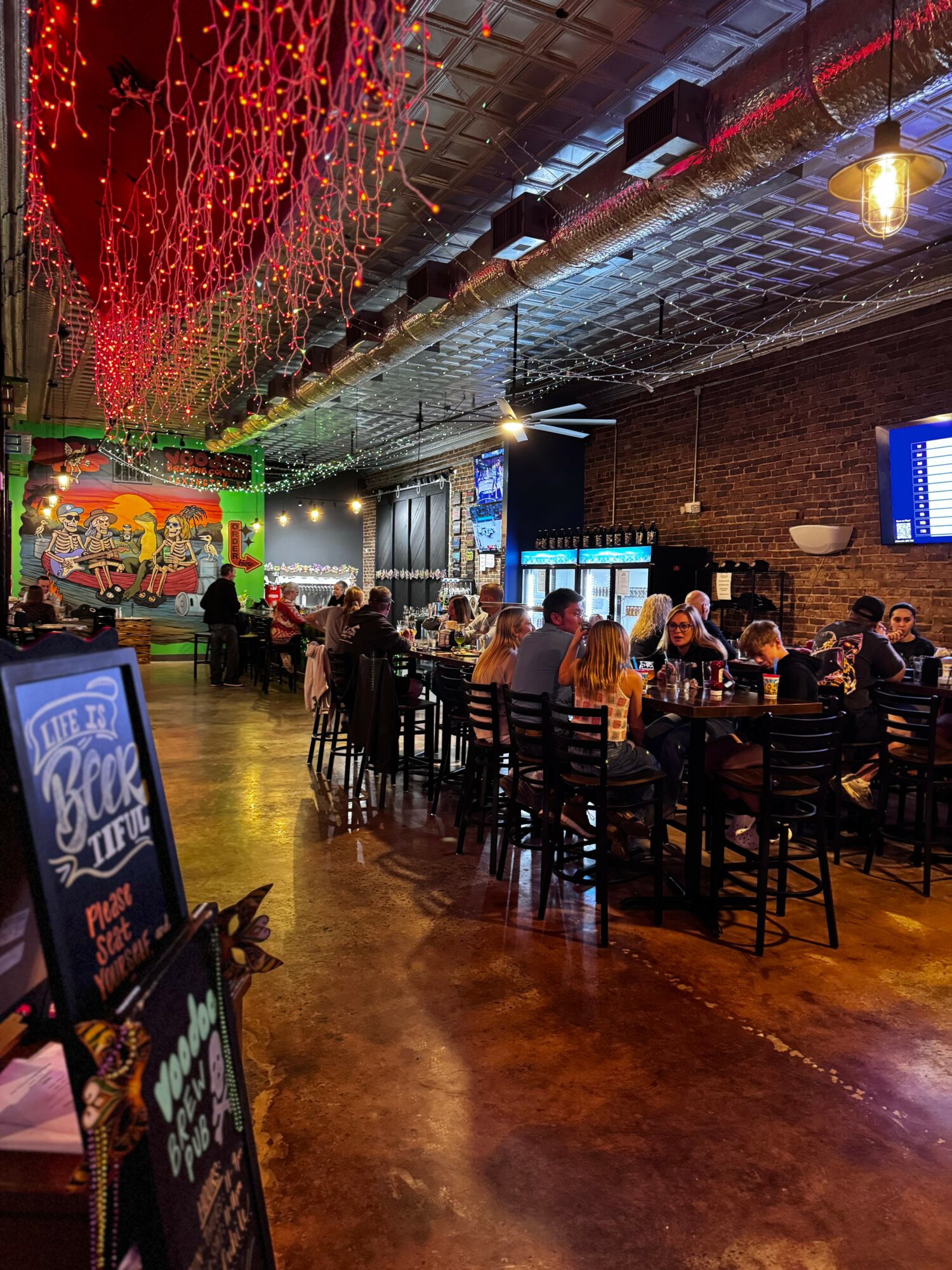 Indoor bar with people sitting at tables, colorful wall art, string lights, and a large screen, warm lighting, brick wall.
