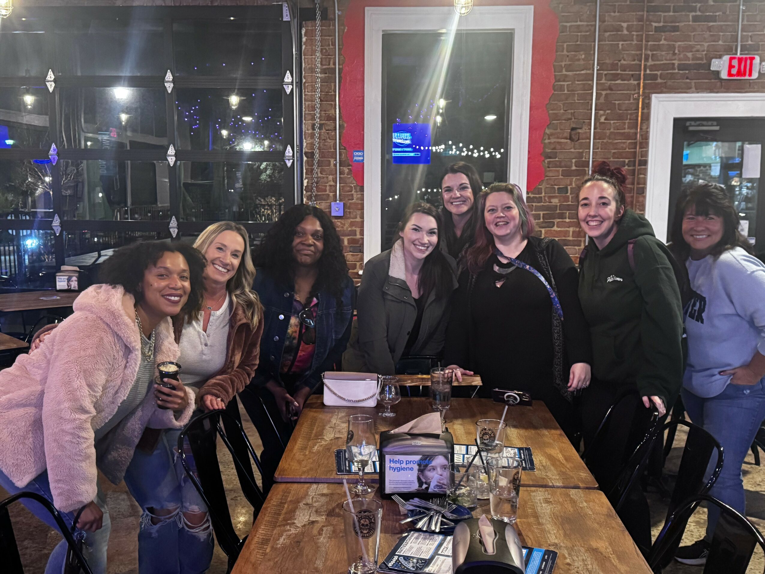 Group of eight women smiling and posing around a table in a restaurant at night, with drinks and a photo frame on the table.