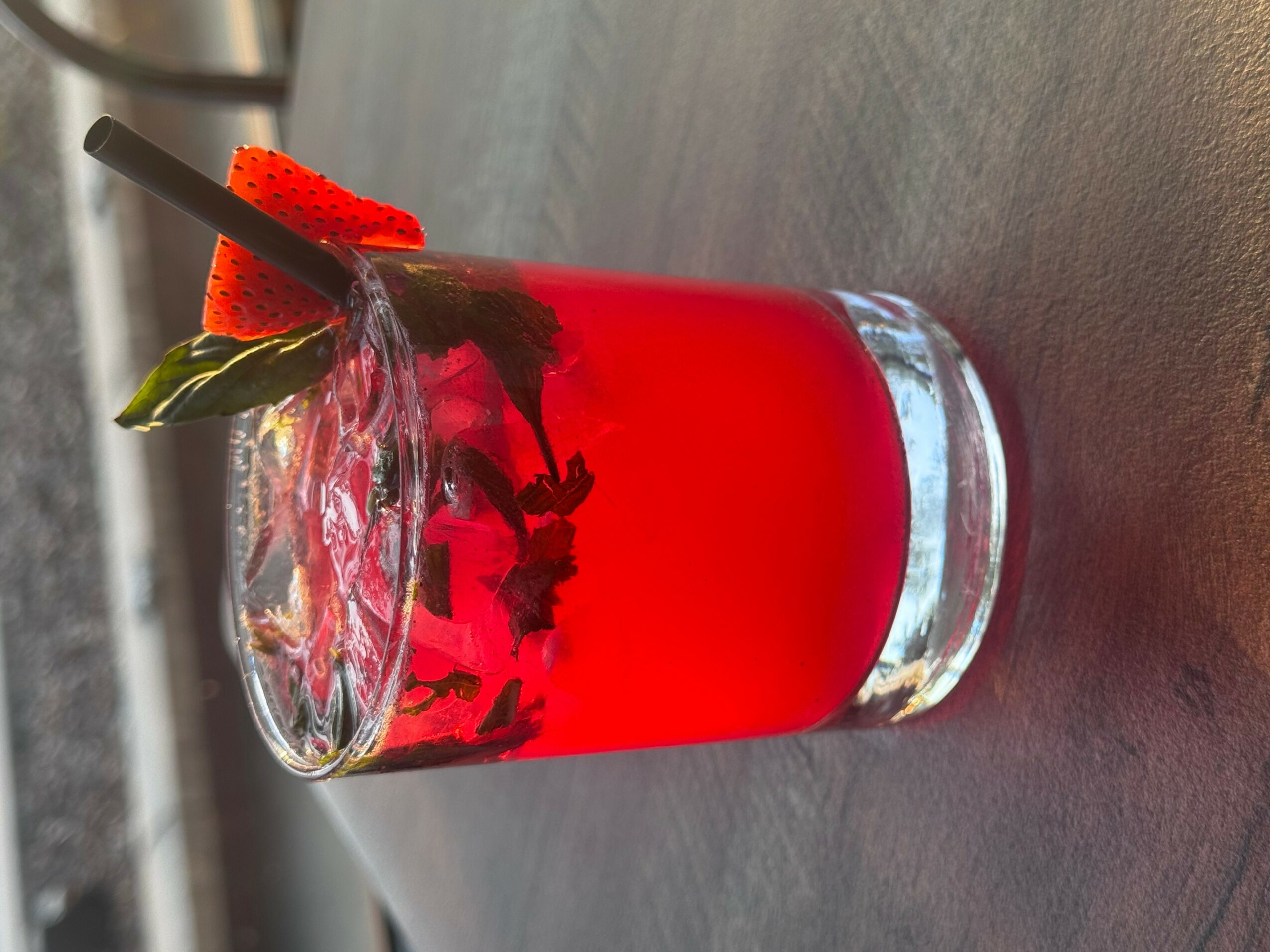 Red cocktail in a glass with a strawberry, mint, and a straw, on a gray surface.