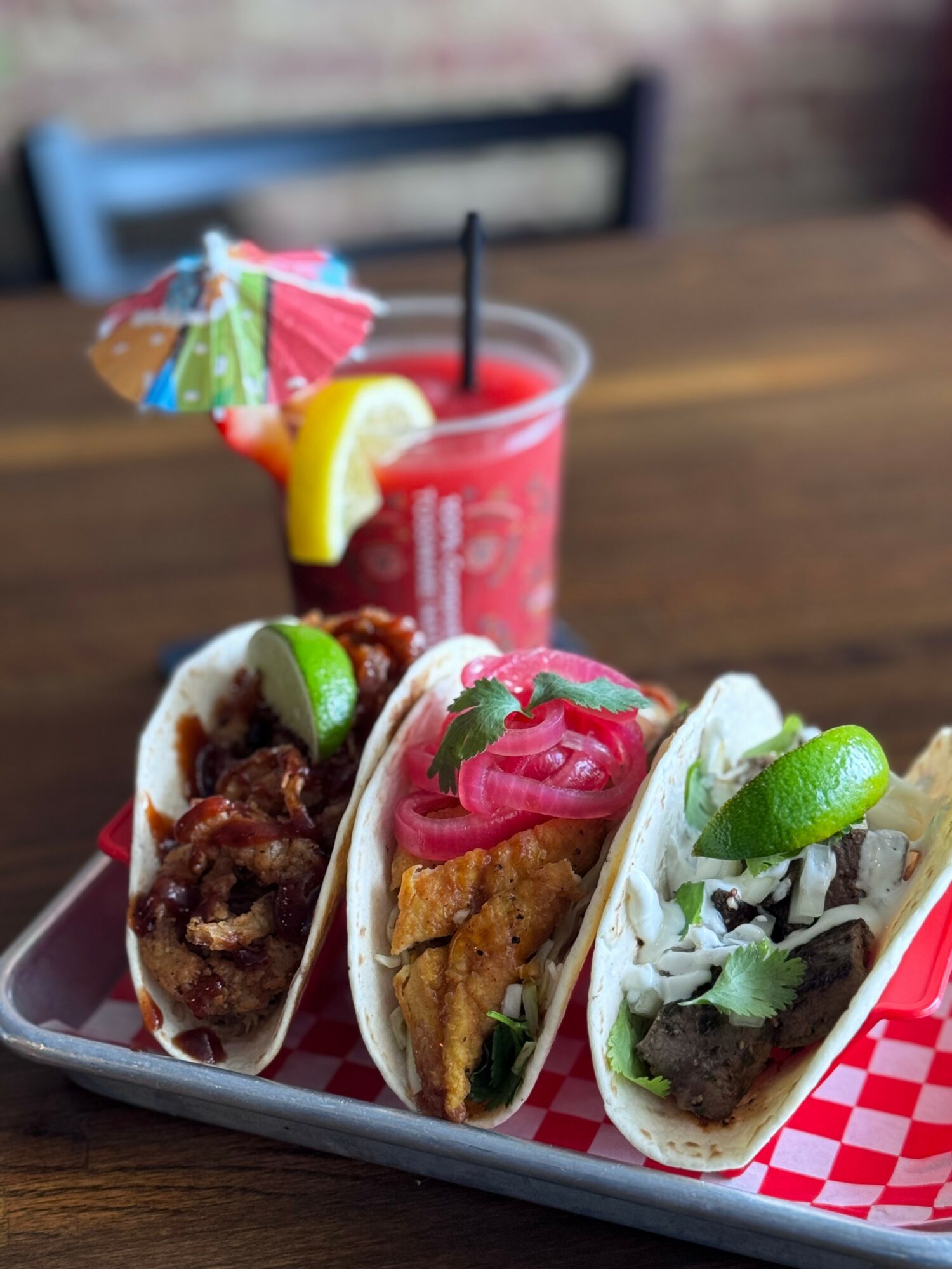 Three tacos with various fillings on a tray, with a pink drink and umbrella in the background.
