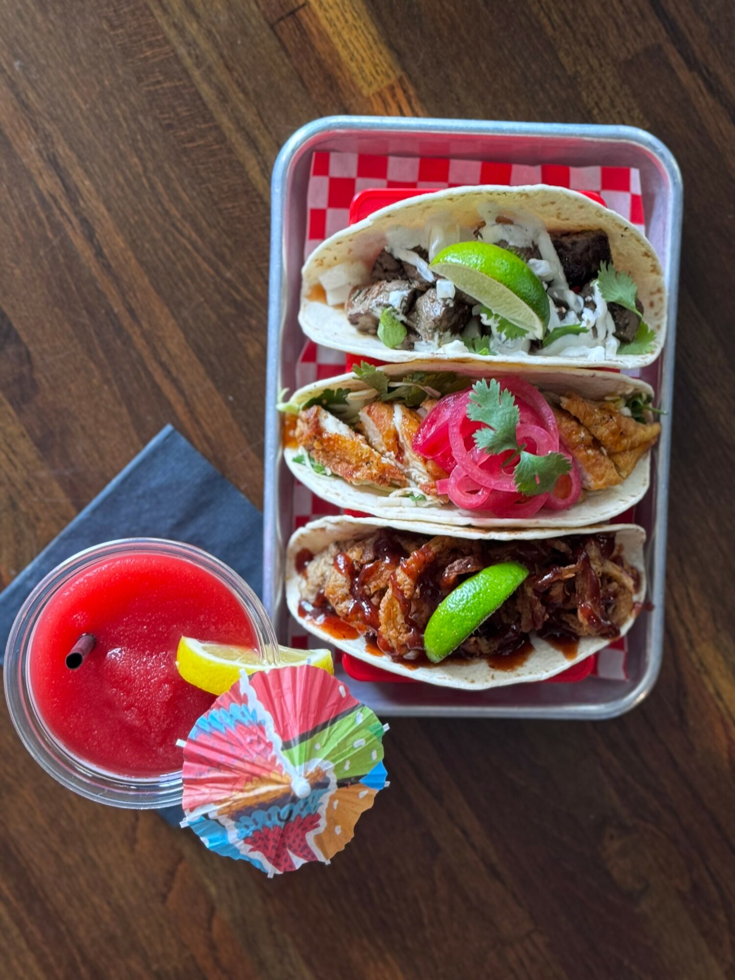 Three tacos in a container with lime, onion, and cilantro, next to a red drink with a colorful umbrella, on a wooden table.