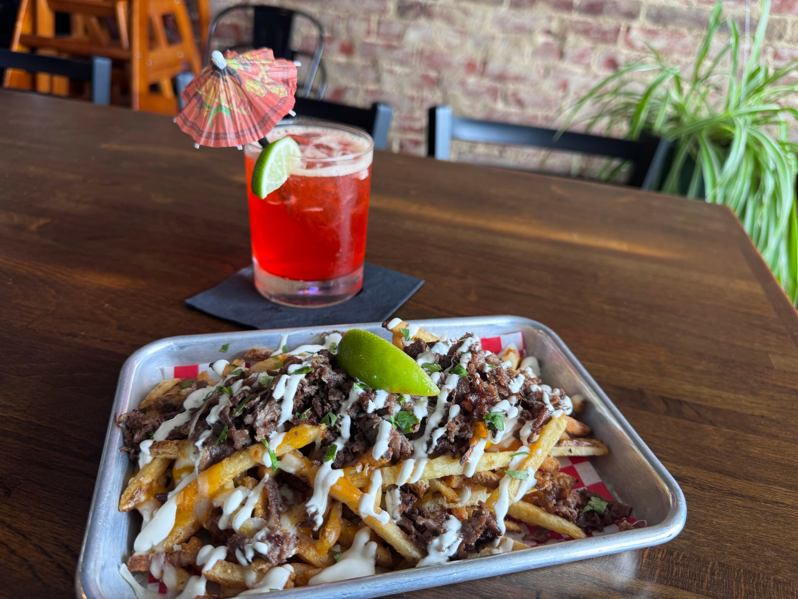 Tray of loaded fries with meat, cheese, and toppings, garnished with lime wedge, on a wooden table, with a red drink in the background.