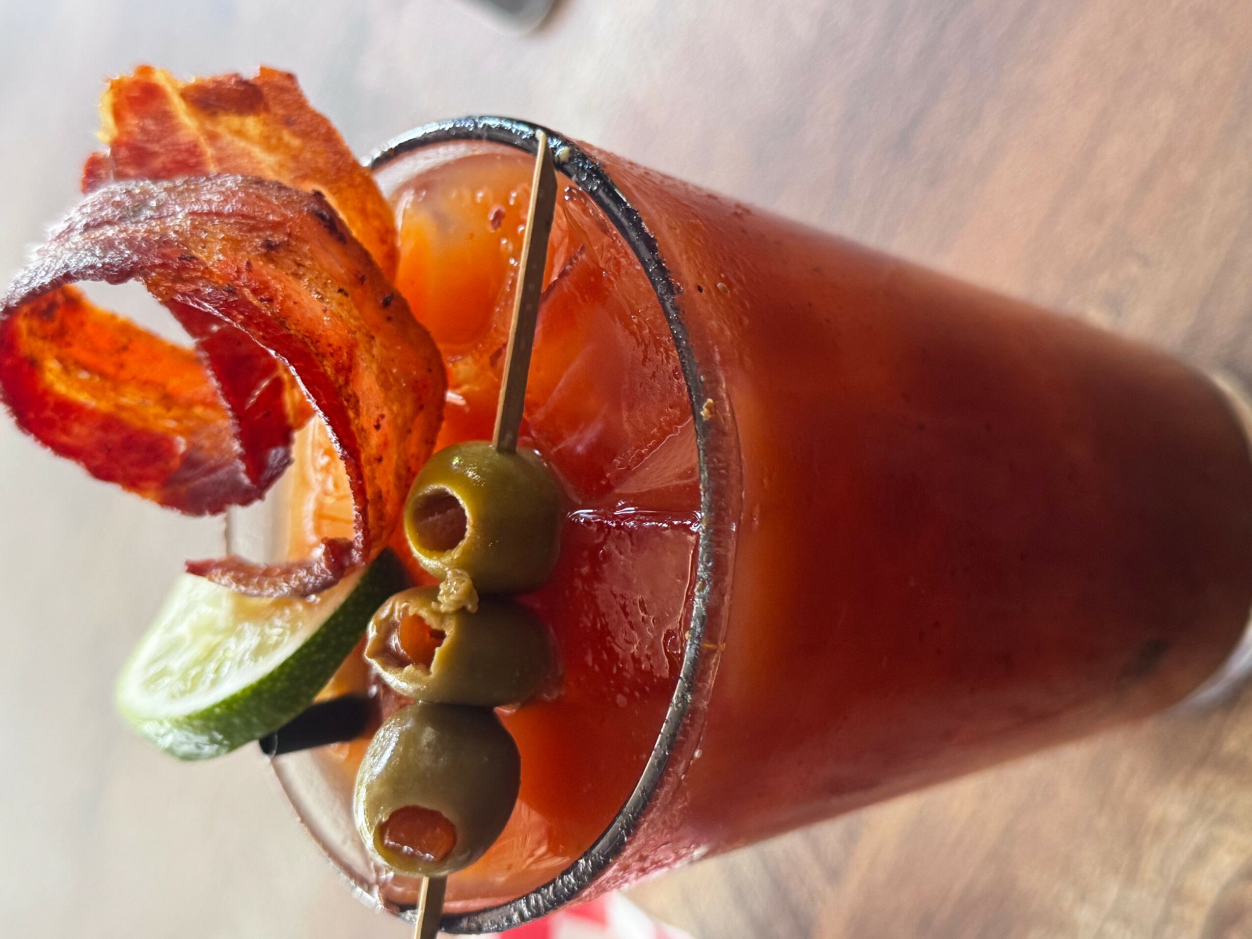 A Bloody Mary cocktail garnished with bacon, lime, and green olives, in a tall glass on a wooden surface.