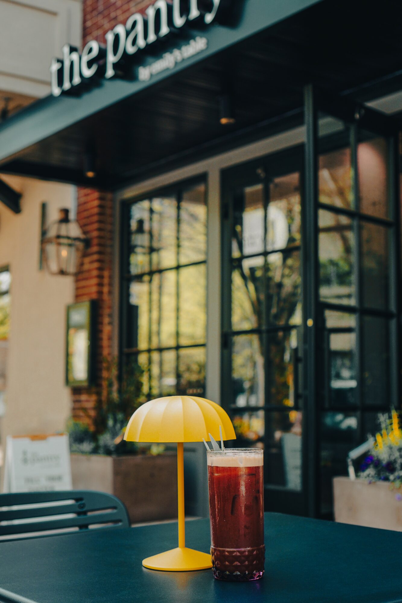 Yellow lamp and a tall glass of red beverage on a dark table outside a cafe with large windows and trees reflected in glass.