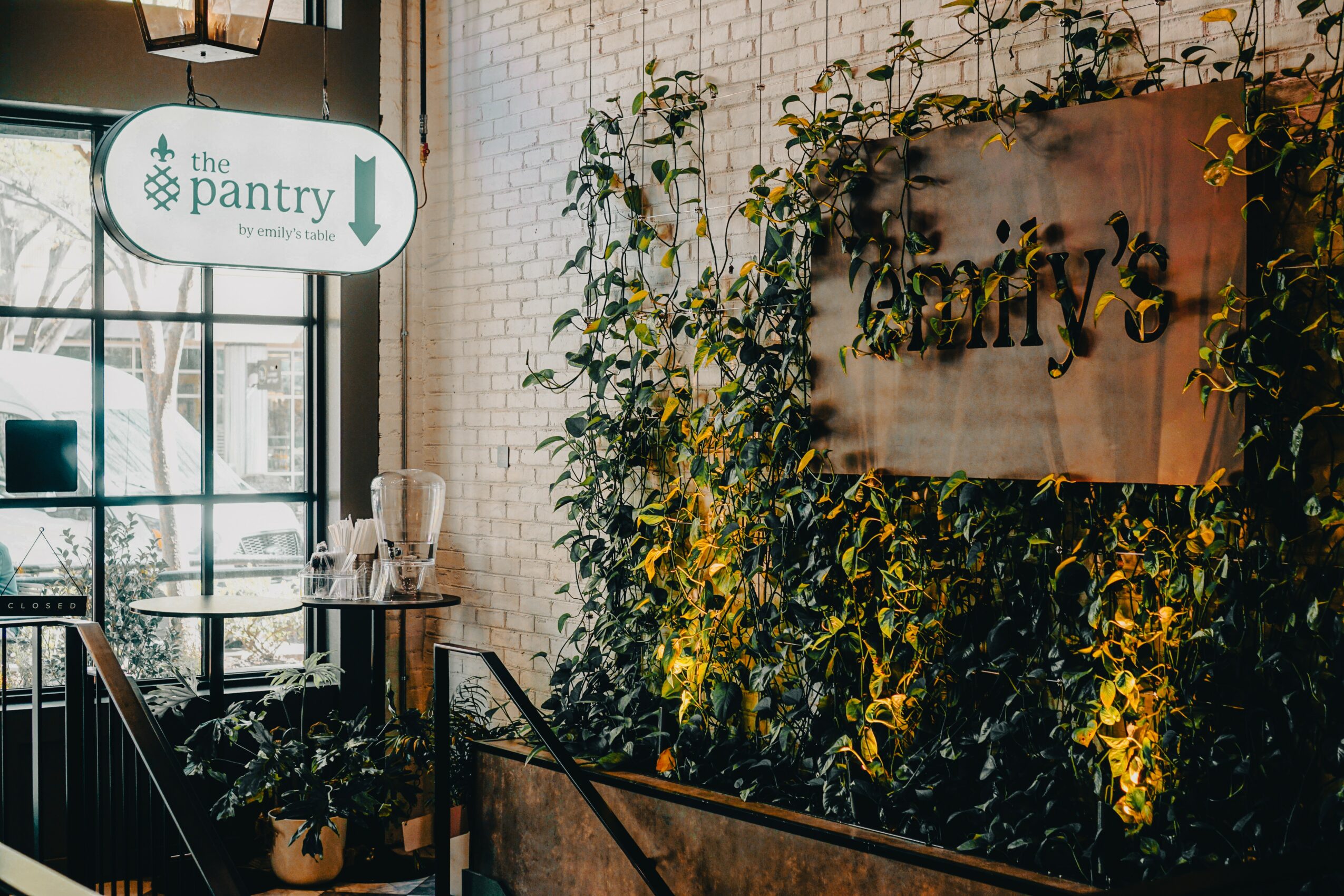 Interior of a cafe with a sign reading 'the pantry' and a wall covered in green plants, with a window and table visible.
