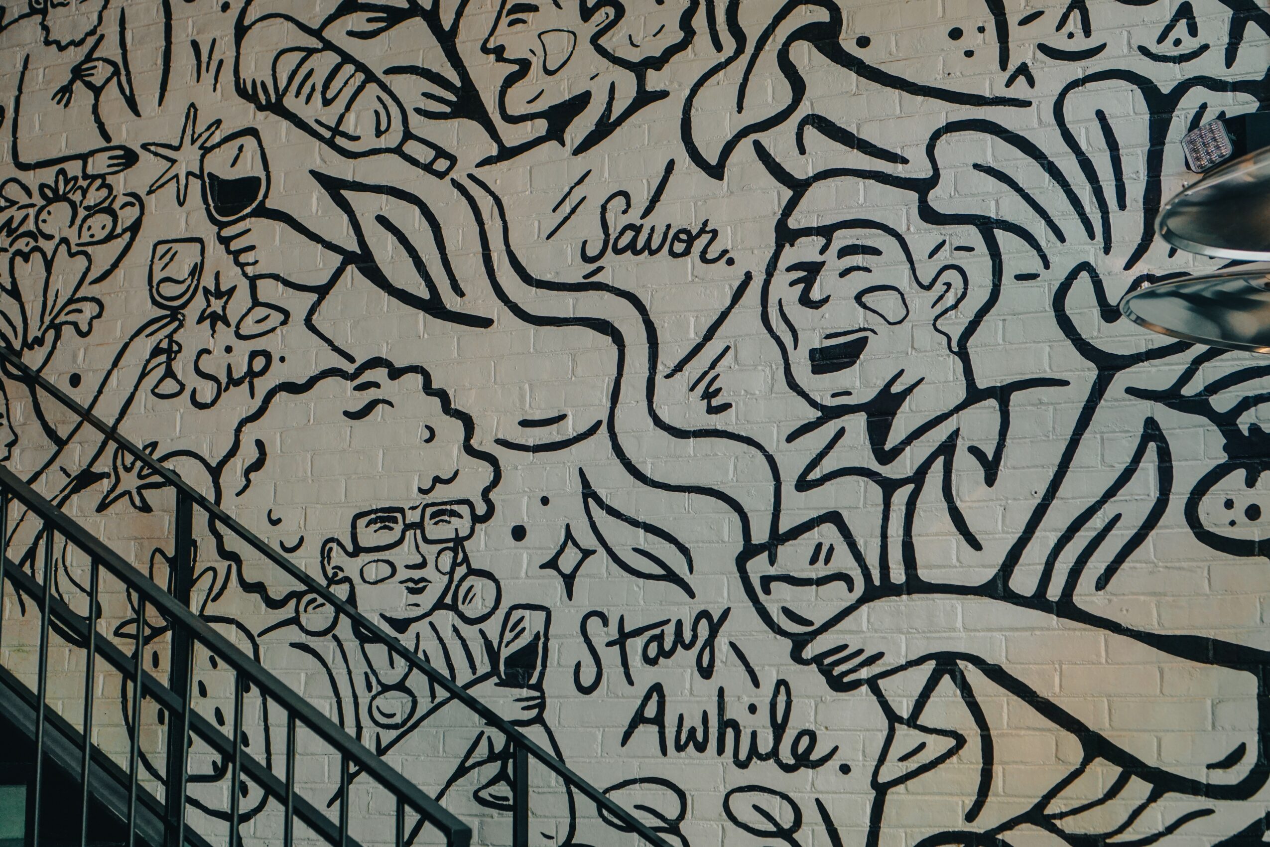 Black and white mural with people holding glasses, a woman with glasses, and text saying 'Savor,' 'Sip,' and 'Stay Awhile.'