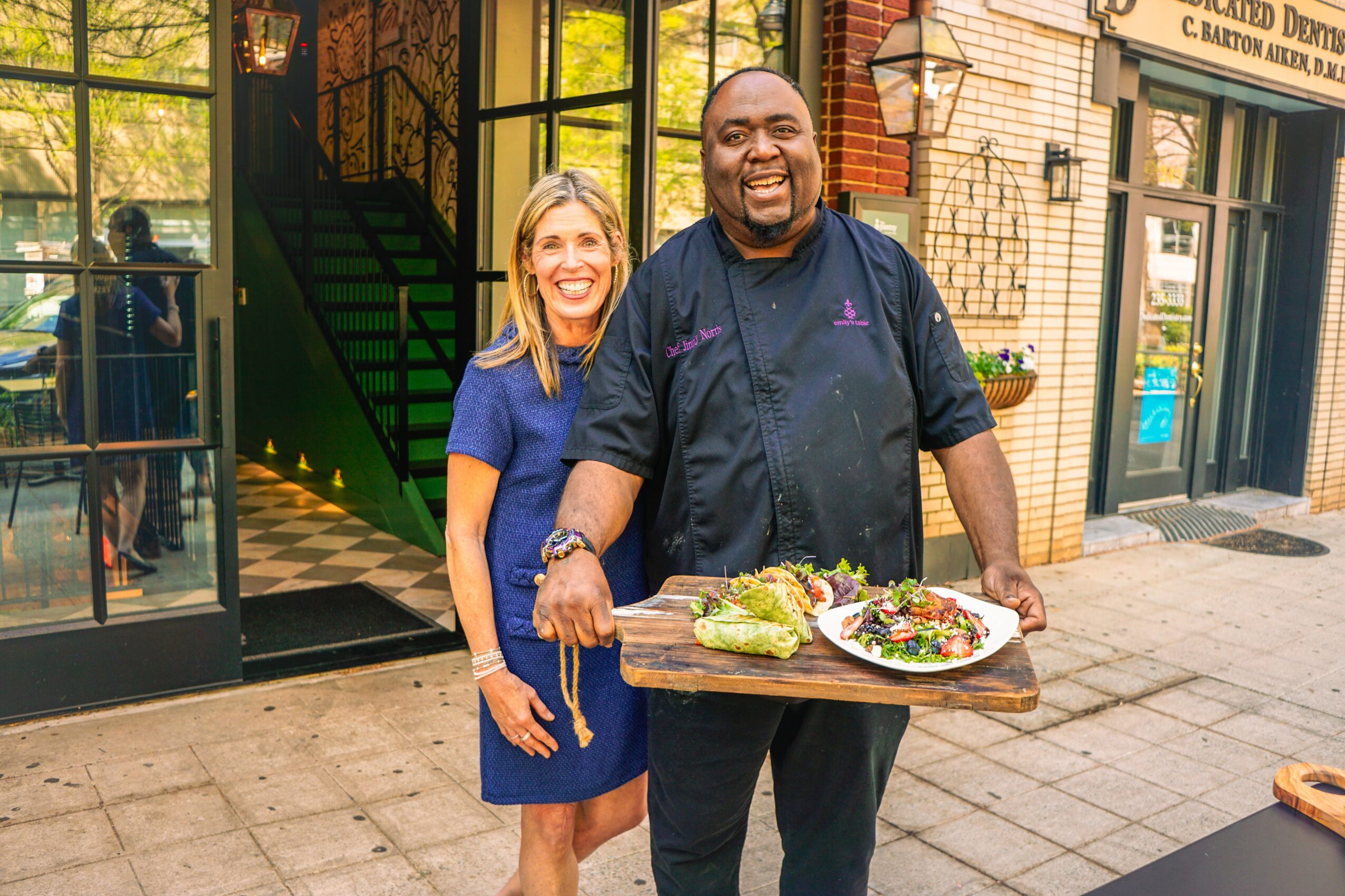 Two smiling people, a woman and a chef, standing outside a restaurant with a tray of food, on a city sidewalk.