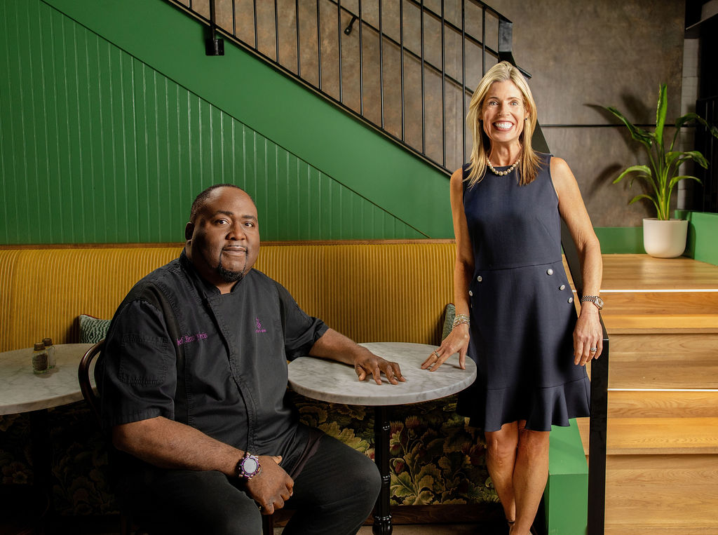 A woman in a navy dress and a man in a black chef's coat sit and stand at a table in a modern interior with green and yellow walls.