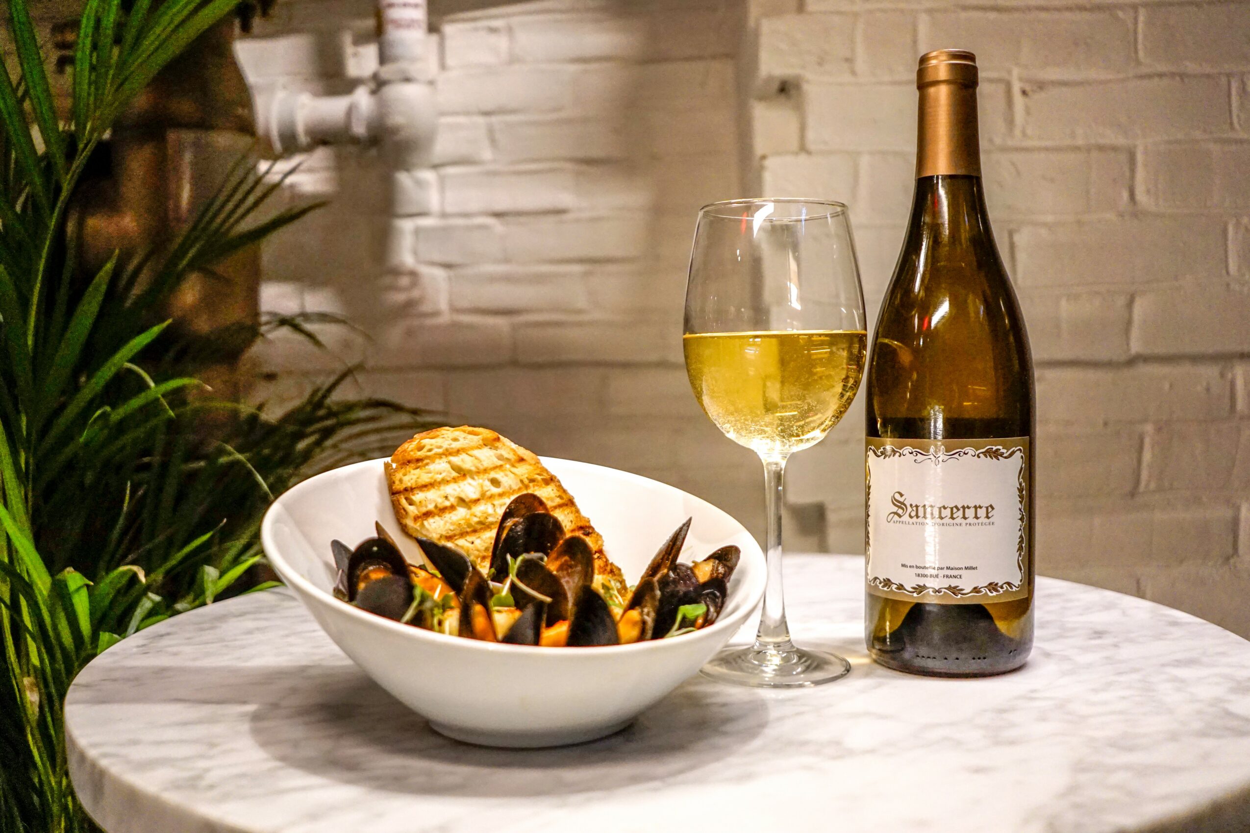 A bowl of mussels and grilled bread on a table with a glass of white wine and a wine bottle, against a brick wall.