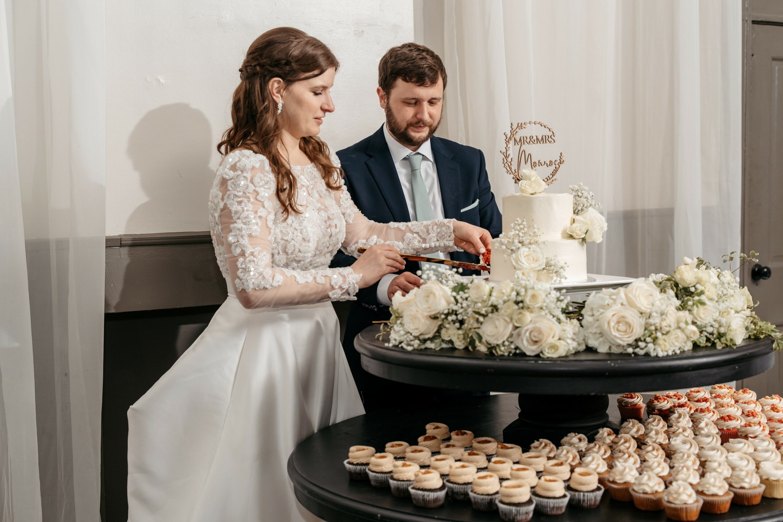 Bride and groom cutting wedding cake with flowers and cupcakes on table, indoor setting.
