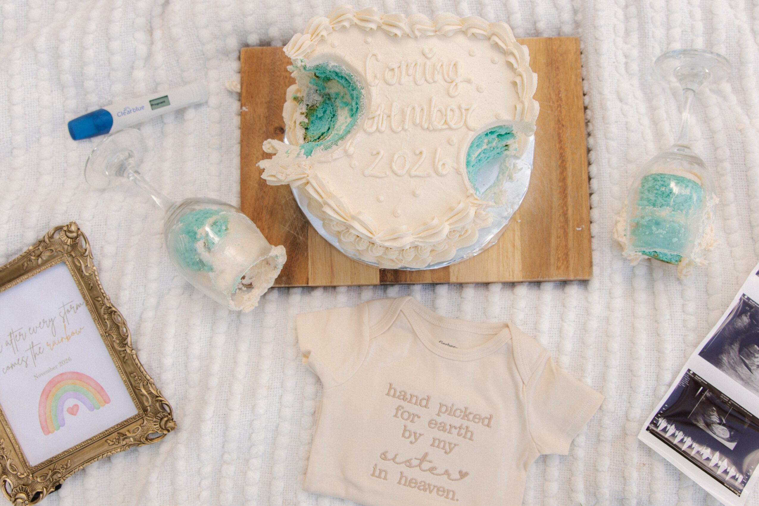 Round cake with white frosting and teal decorations, a small shirt, and birthday items on a white textured surface.