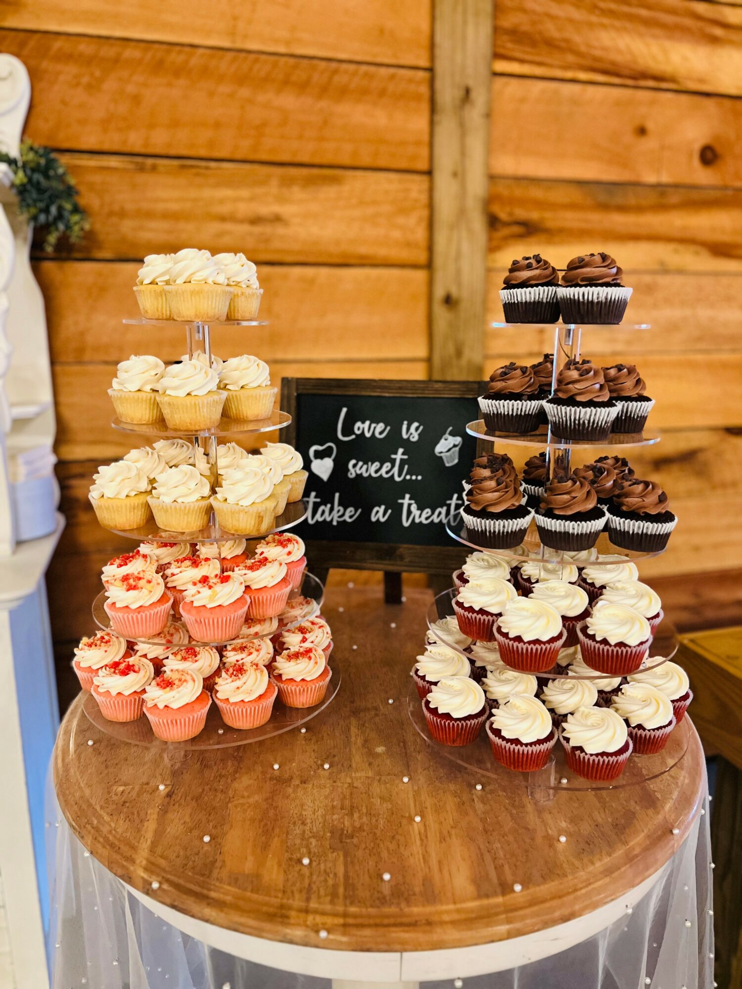 Assorted cupcakes on a wooden table with a wooden wall background, decorated with a small blackboard sign, and a white cloth underneath.