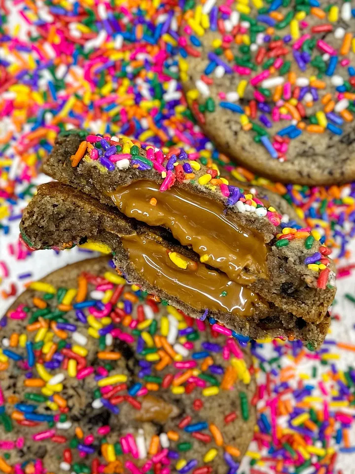 Close-up of cookies with colorful sprinkles and caramel filling, with one broken cookie revealing caramel inside, on a sprinkle-covered surface.