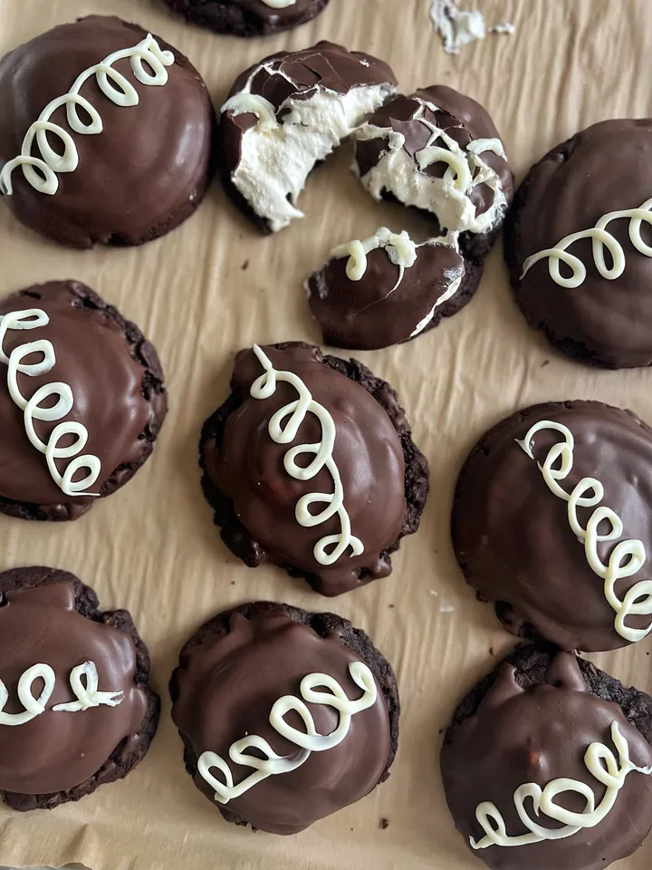 Chocolate-covered cookies with white icing swirl decoration on a wooden surface, one cookie broken revealing filling inside.