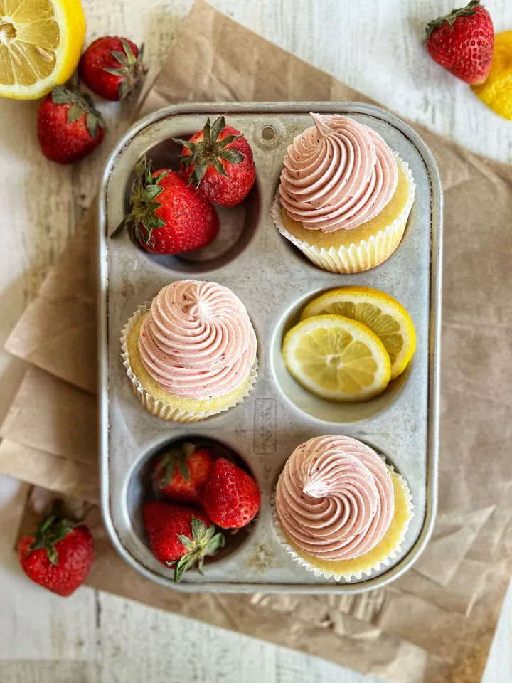 Three cupcakes with pink frosting, lemon slices, and fresh strawberries on a metal tray, with strawberries and lemon slices around.