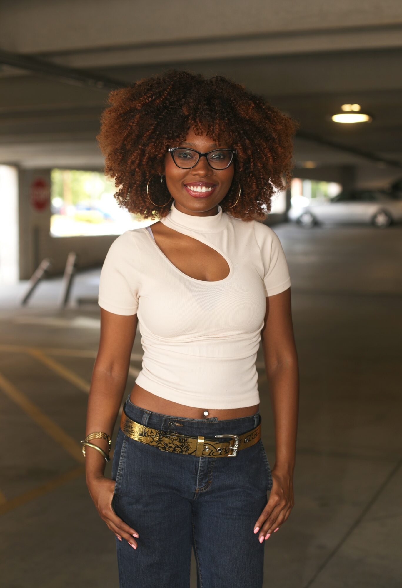 Smiling woman with curly hair and glasses standing in a parking garage, wearing a white top and jeans.