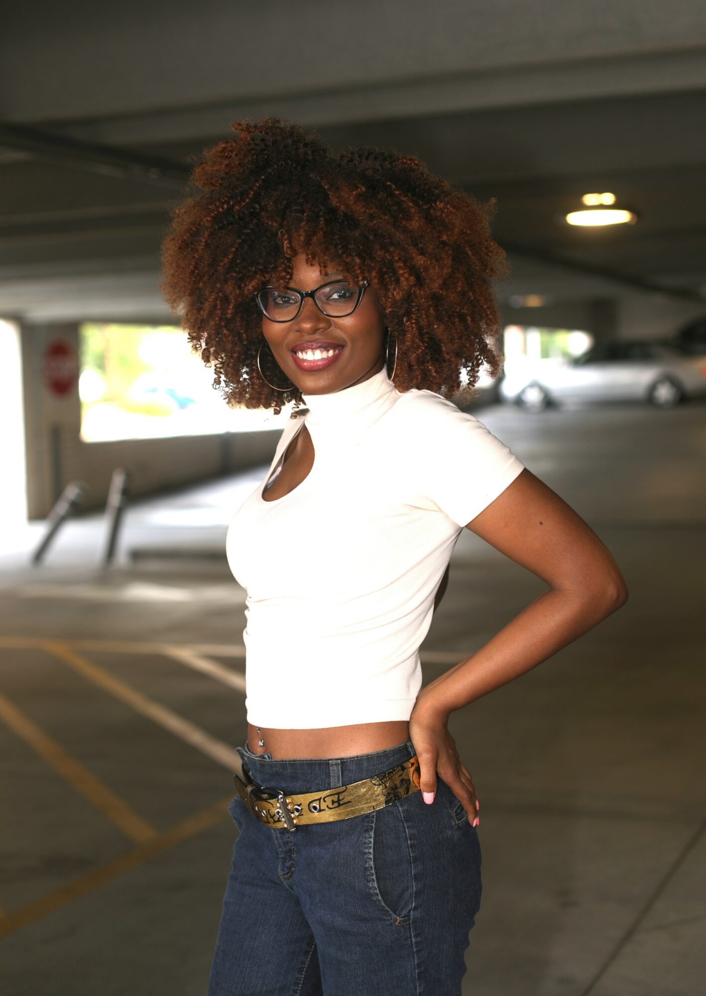 Smiling woman with curly hair and glasses in a parking garage, wearing a white top and jeans, with one hand on her hip.