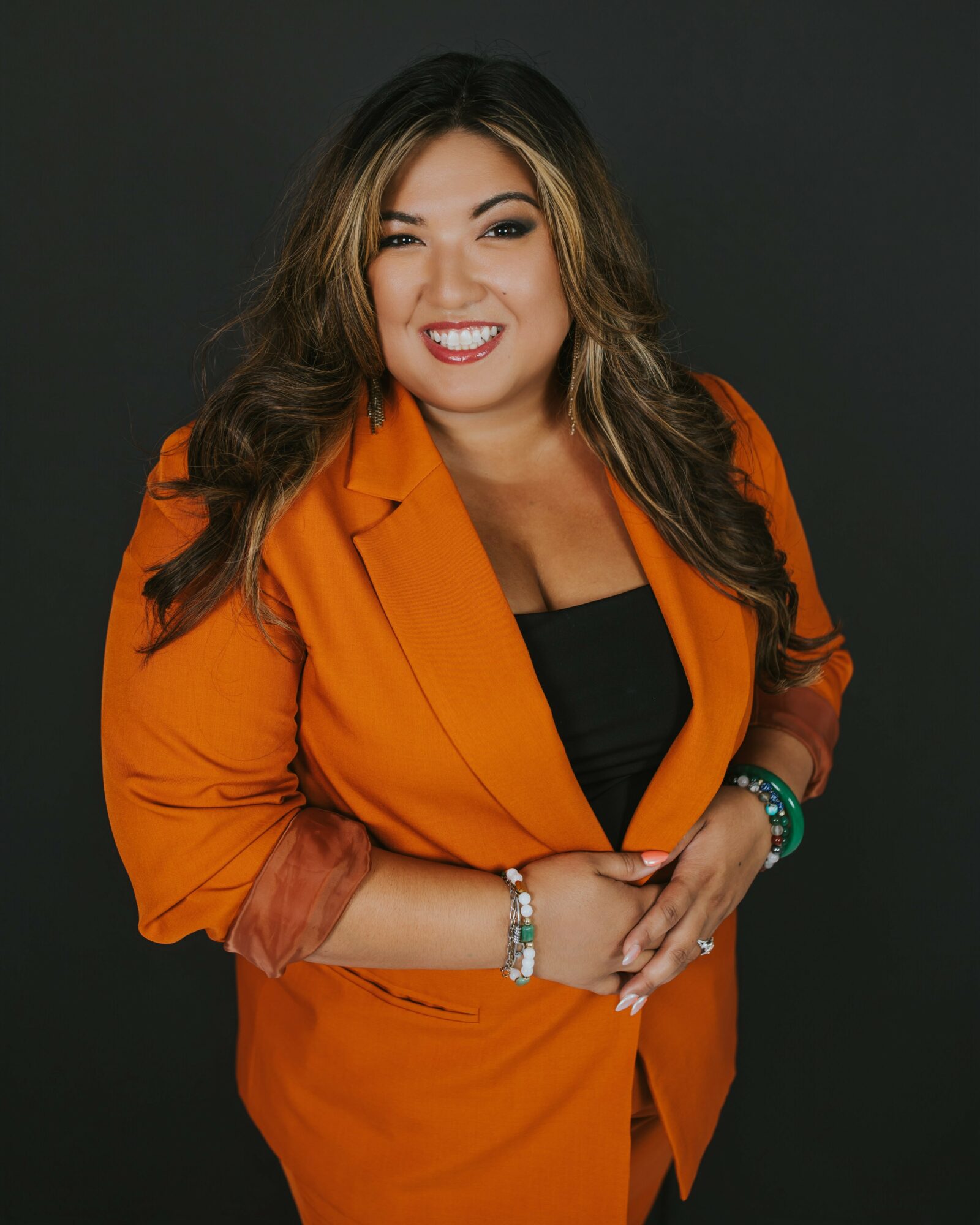 Woman with long wavy hair smiling, wearing an orange blazer over a black top, against a dark background.