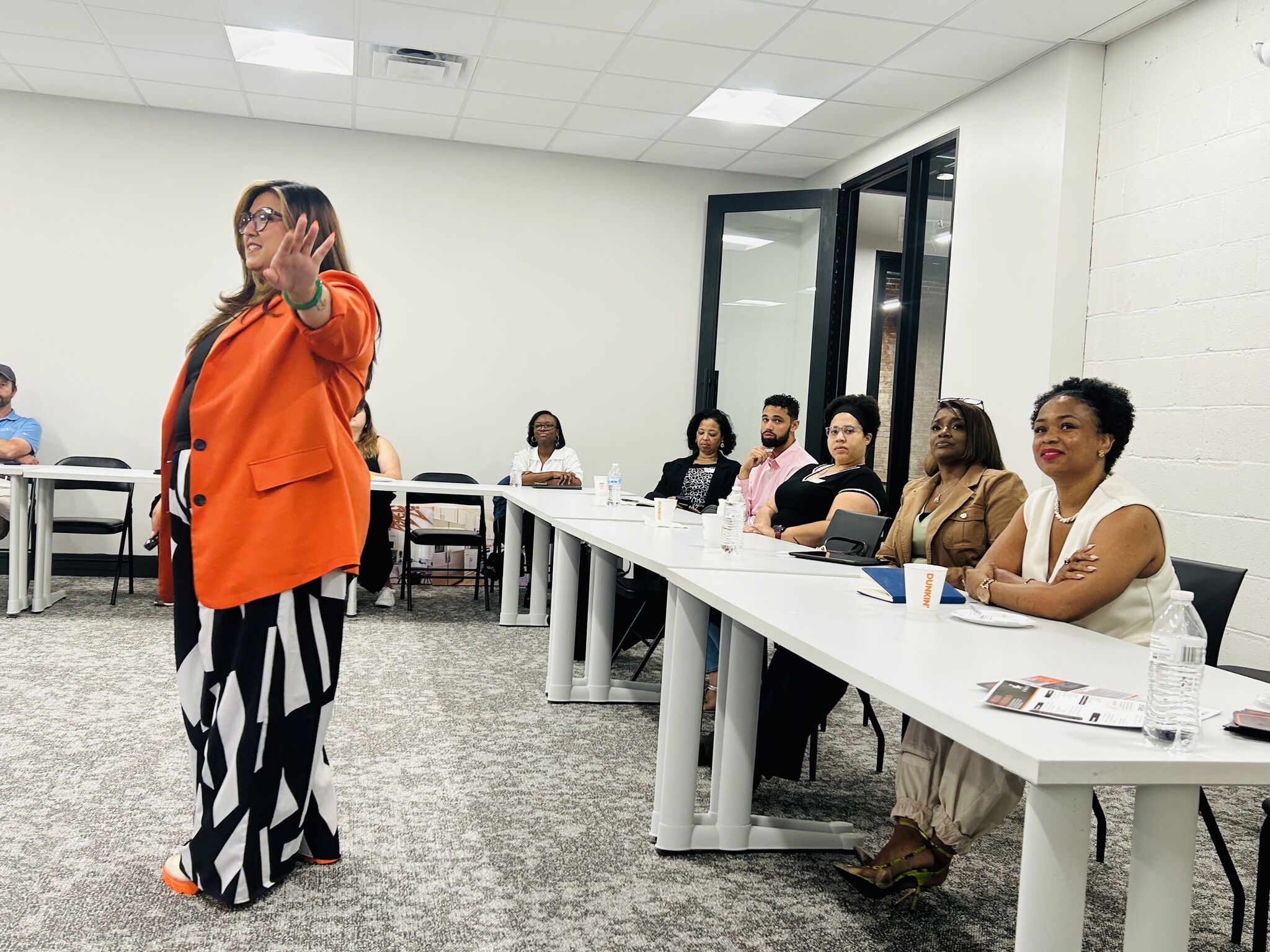 Woman in orange blazer speaking to seated group in a conference room, some taking notes, others listening attentively.