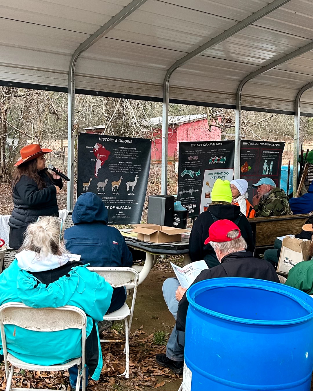 Group of people seated outdoors under a shelter, listening to a woman speaking with informational posters behind her.