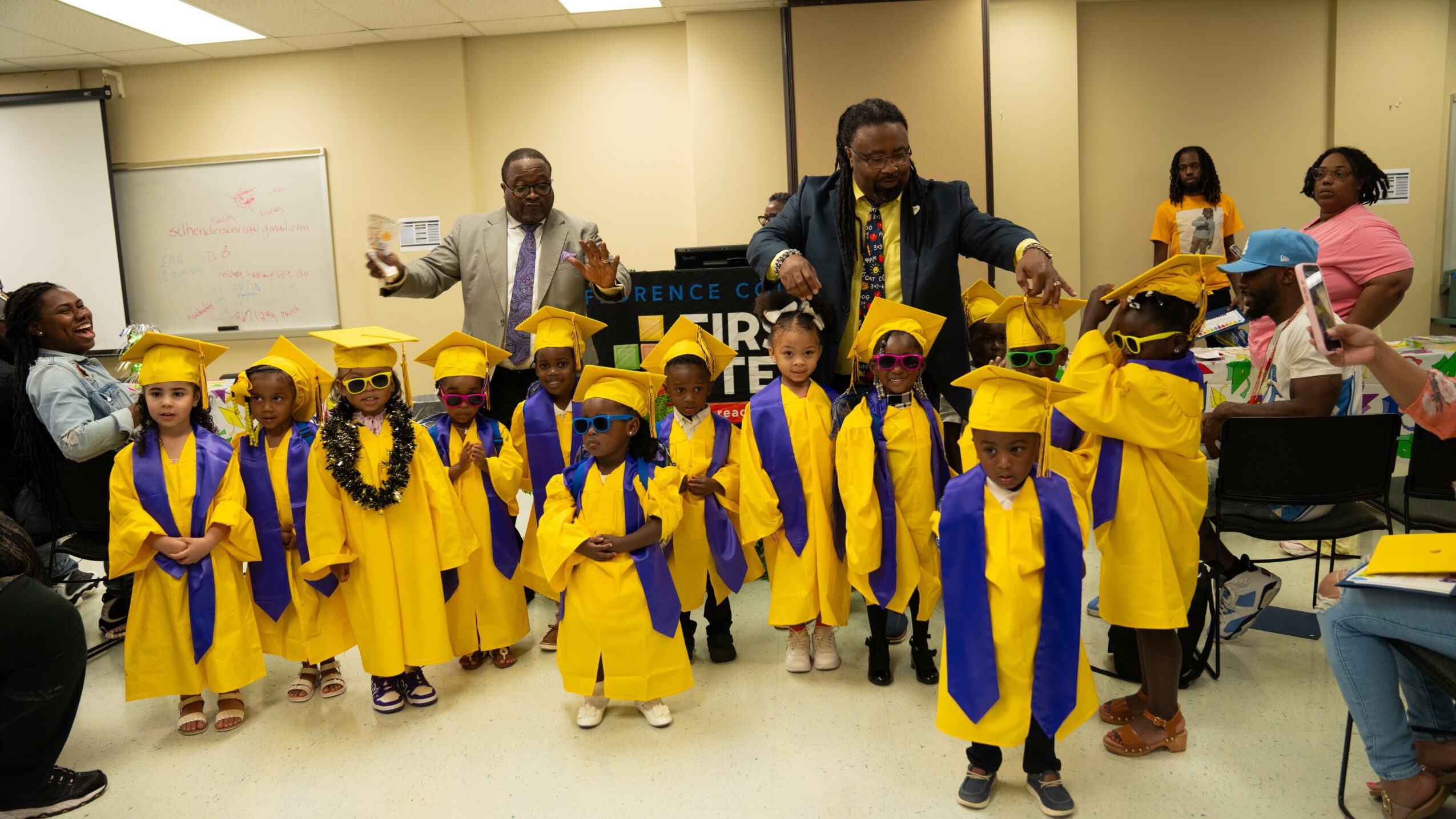 Children in yellow caps and gowns stand in a classroom, with adults assisting and celebrating graduation.