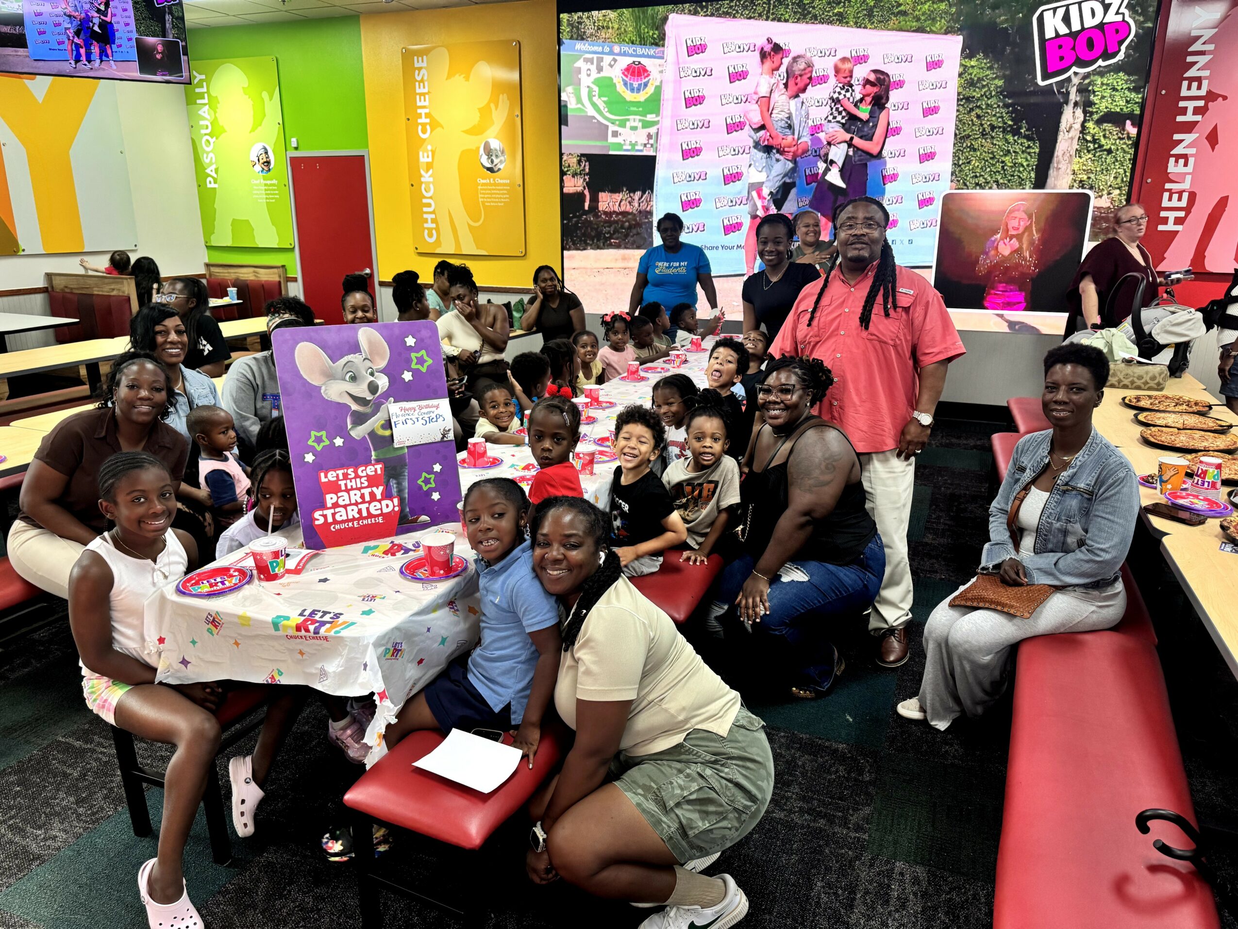 Group of children and adults gathered around a table with colorful decorations in an indoor play area.