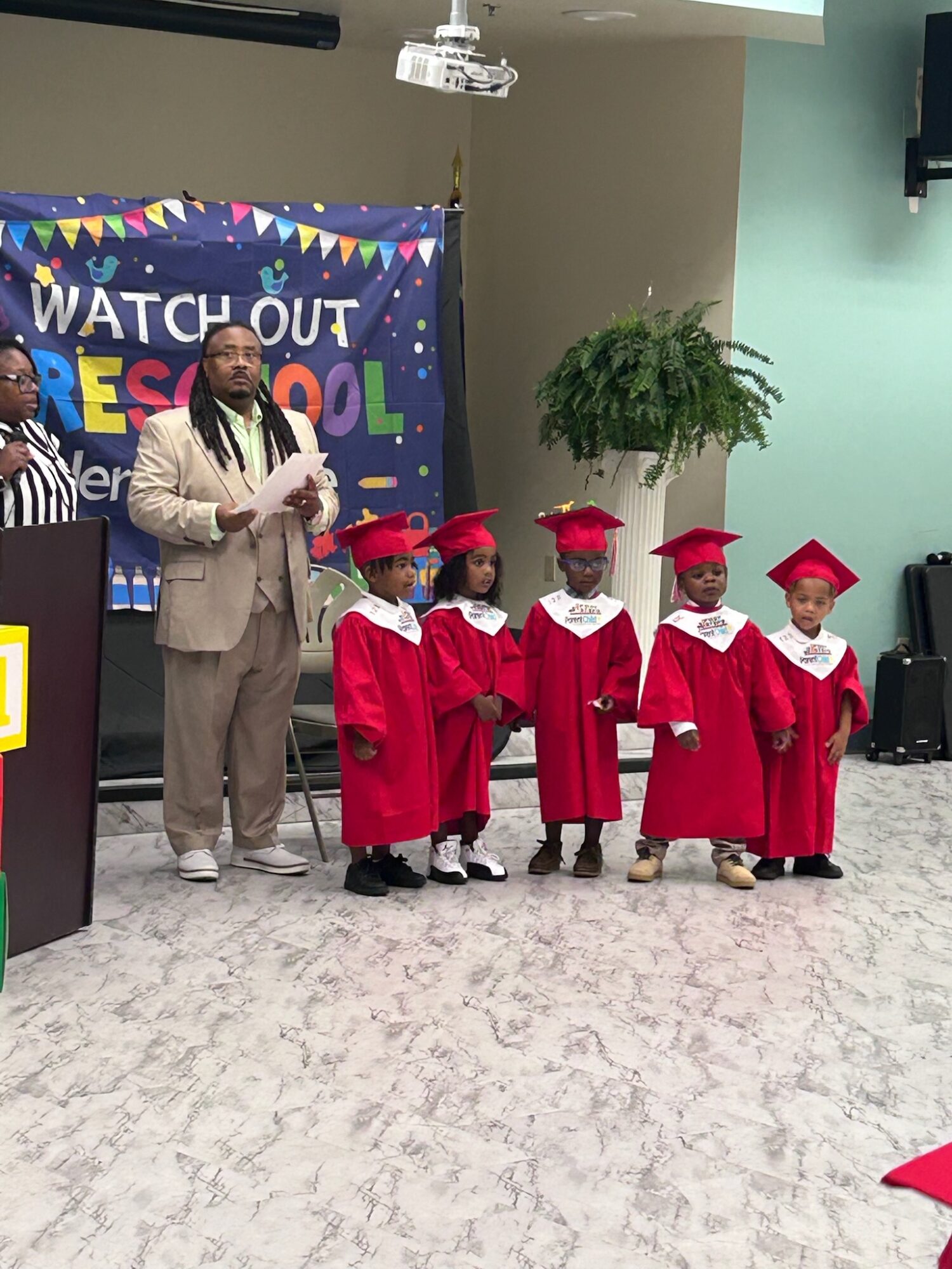Group of young children in red graduation gowns and caps standing on stage, with a woman in a suit and a colorful banner in the background.
