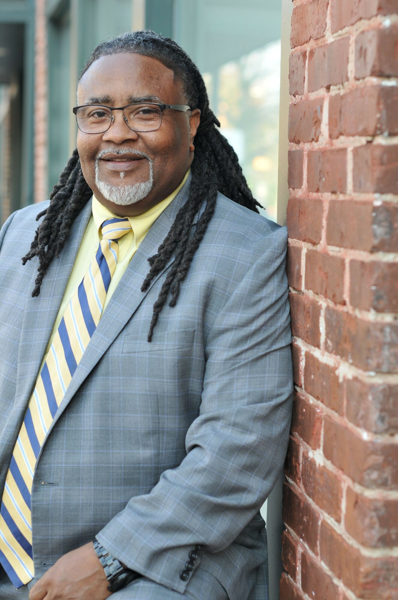 Person with long dreadlocks, glasses, and a suit, smiling outdoors near a brick wall.