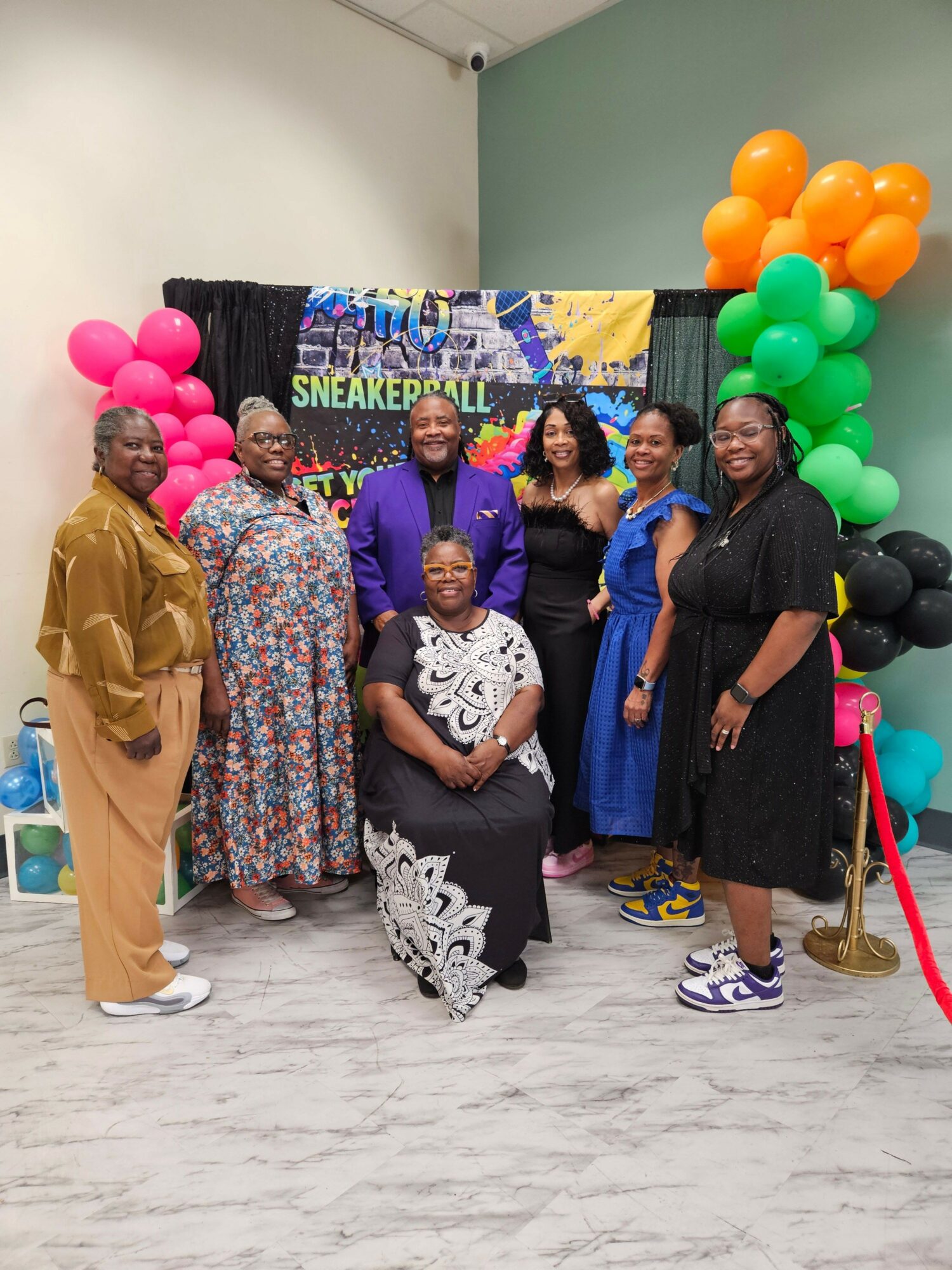 Group of people standing in front of a colorful backdrop with balloons, some holding balloons, indoors.