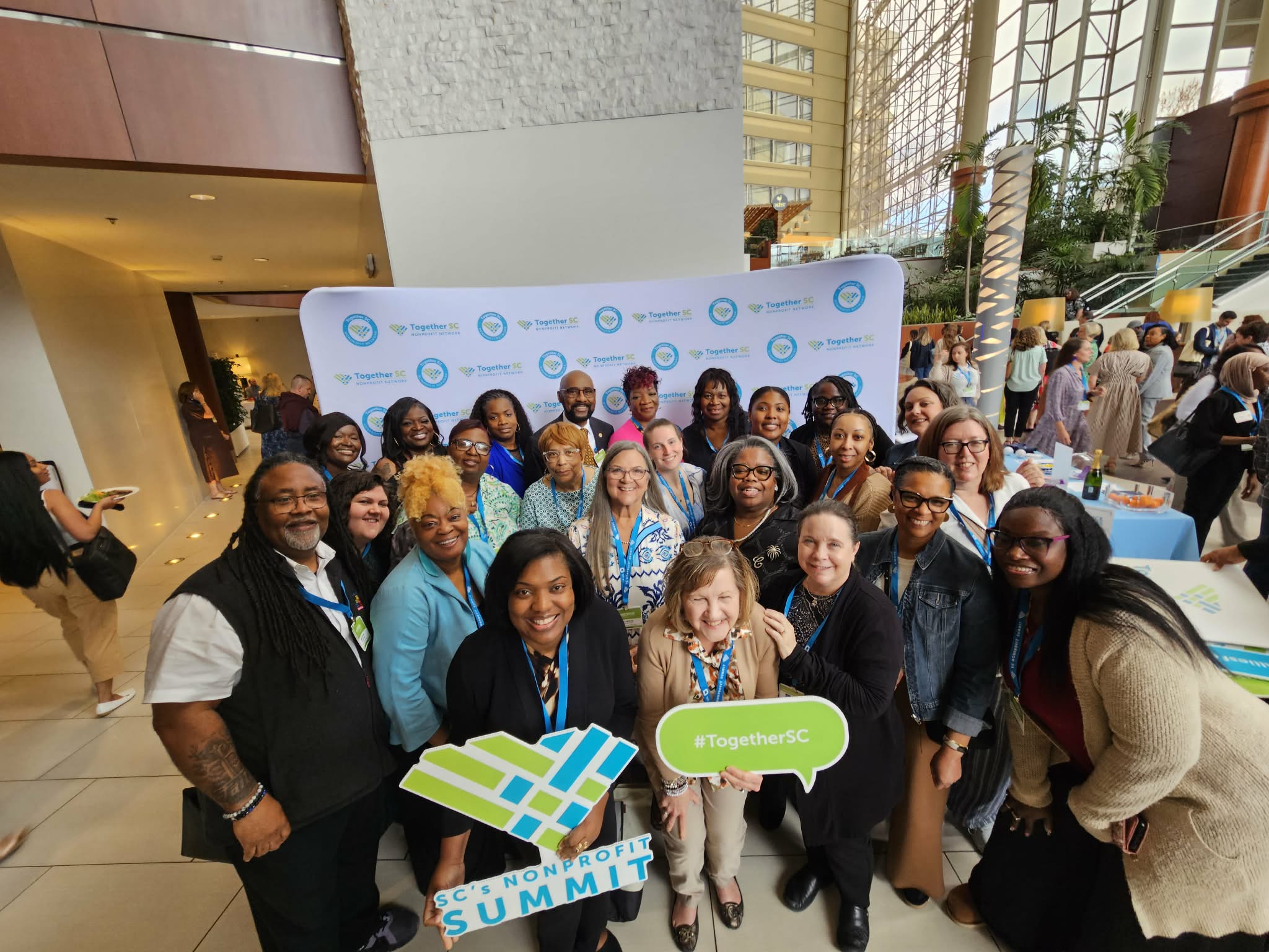 Group of diverse people smiling at a conference or event, standing in front of a backdrop with logos, indoors with large windows.