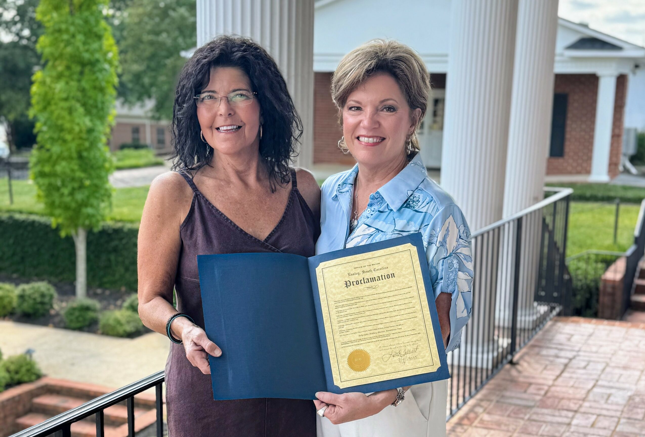 Two women smiling outdoors, holding a certificate in a blue folder, standing on a porch with columns and greenery.