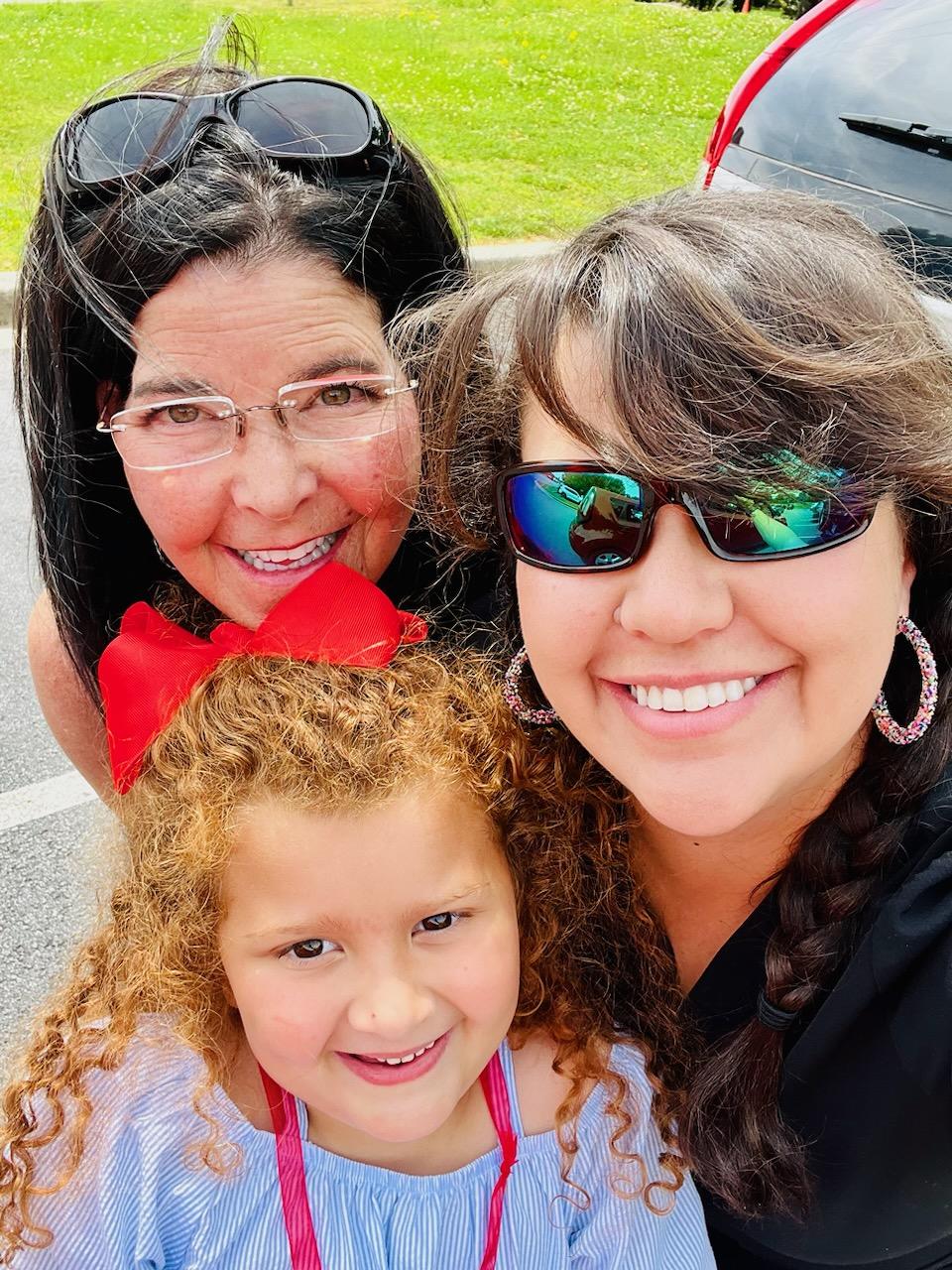 Three smiling females, one child, outdoors with cars in background, all wearing sunglasses and colorful clothing.