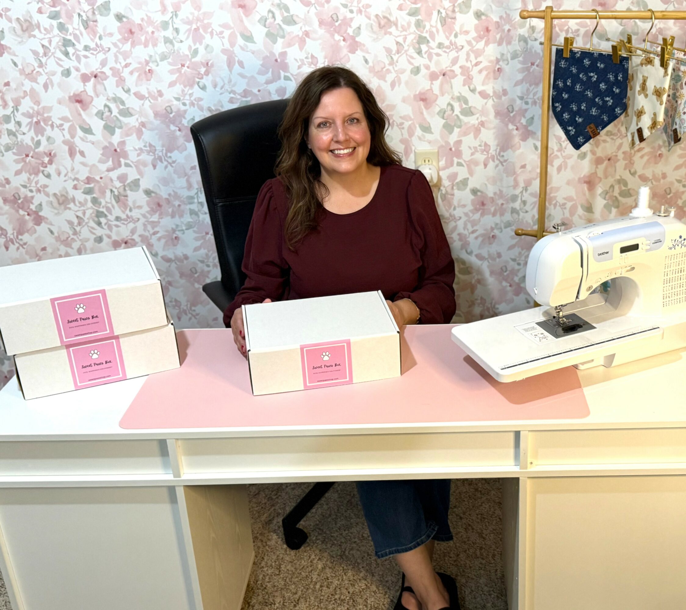 Woman sitting at a desk with sewing machine and boxes, floral wallpaper background, smiling, wearing a maroon top.