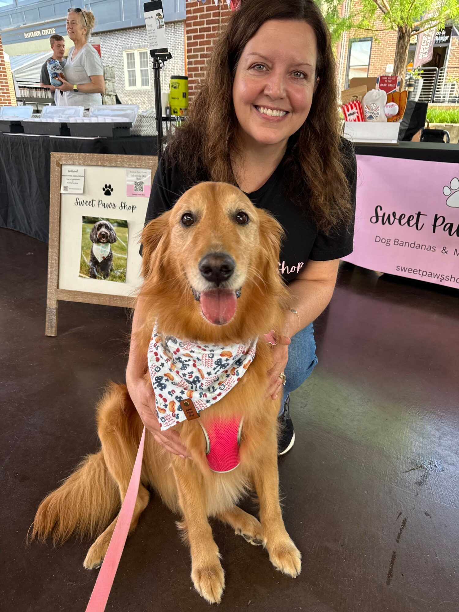 Woman smiling with a golden retriever wearing a bandana, indoors at a table with a pink sign in the background.
