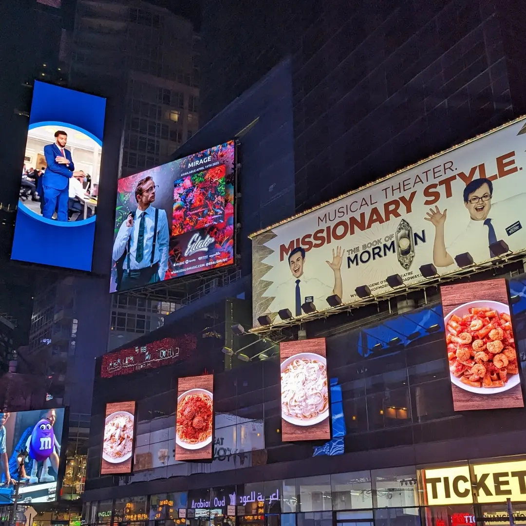 Cityscape with illuminated billboards and advertisements at night, including a musical theater sign and food images.