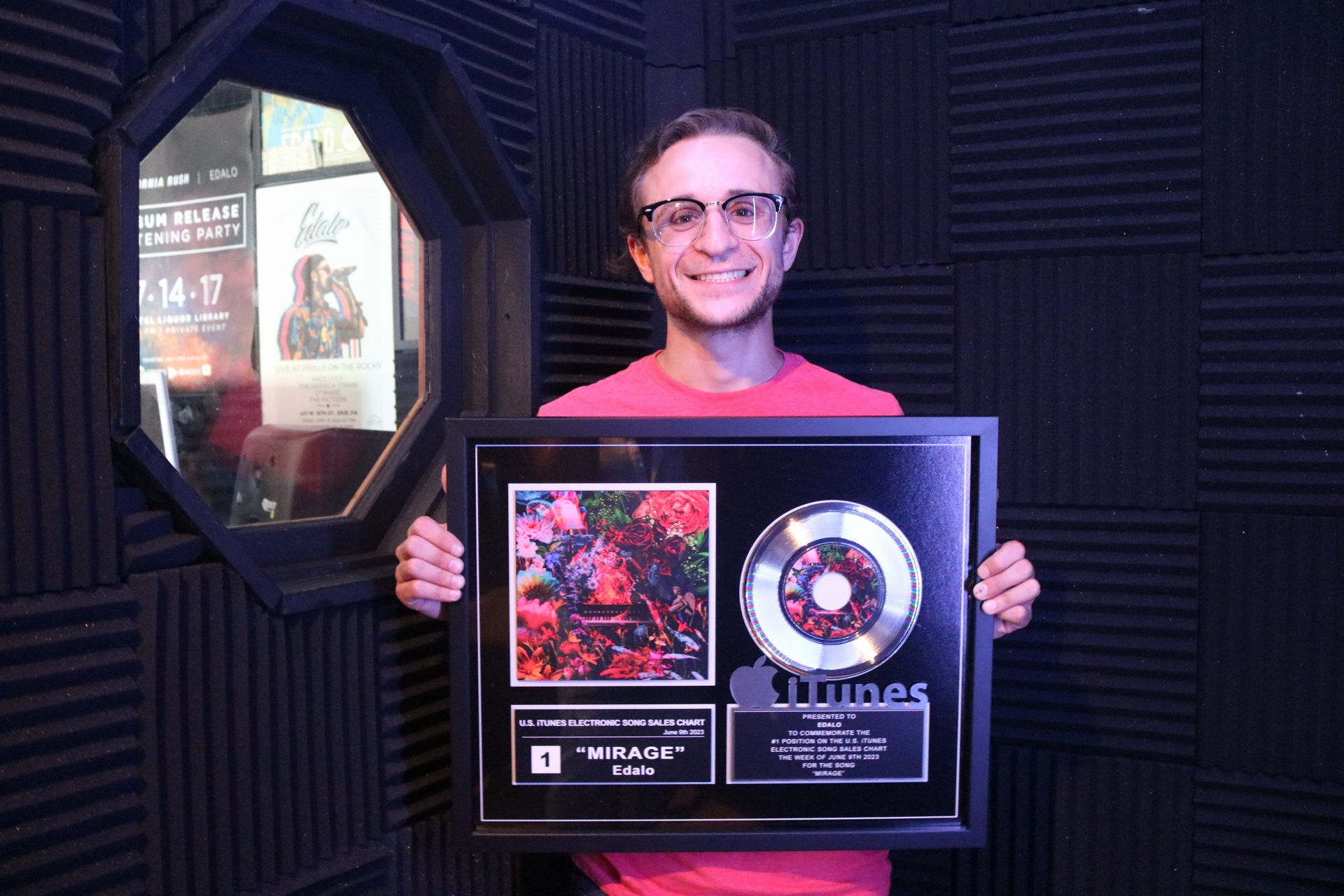 Person with glasses smiling holding a framed music award in a recording studio with black acoustic panels