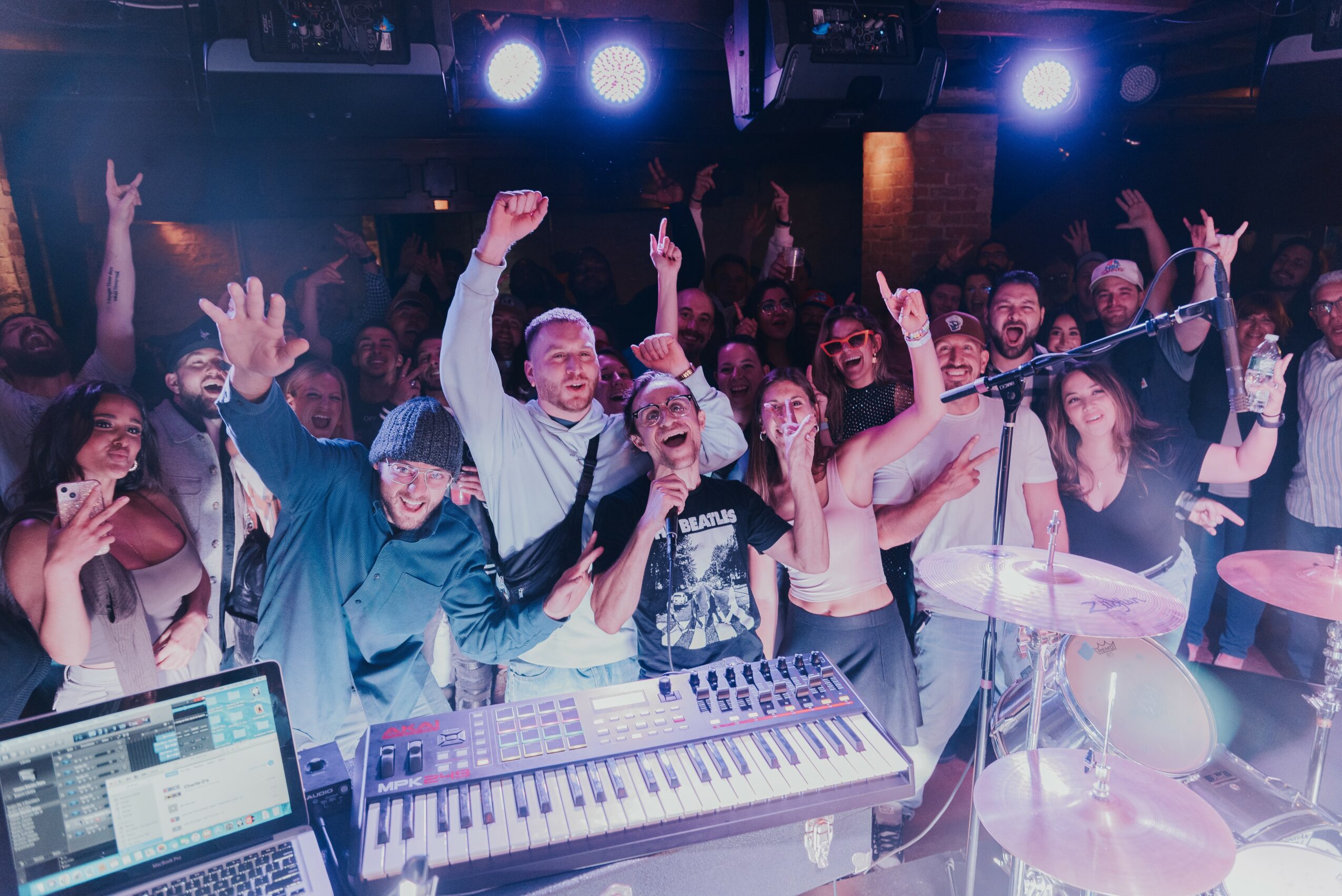Crowd dancing and cheering at a concert with a keyboard and drums in foreground, colorful stage lights overhead.