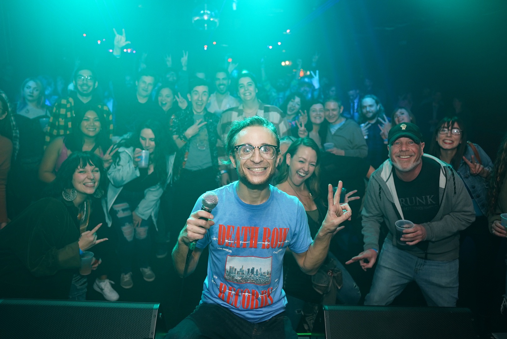 Group of people at a party, smiling and making peace signs, with a man holding a microphone in front.