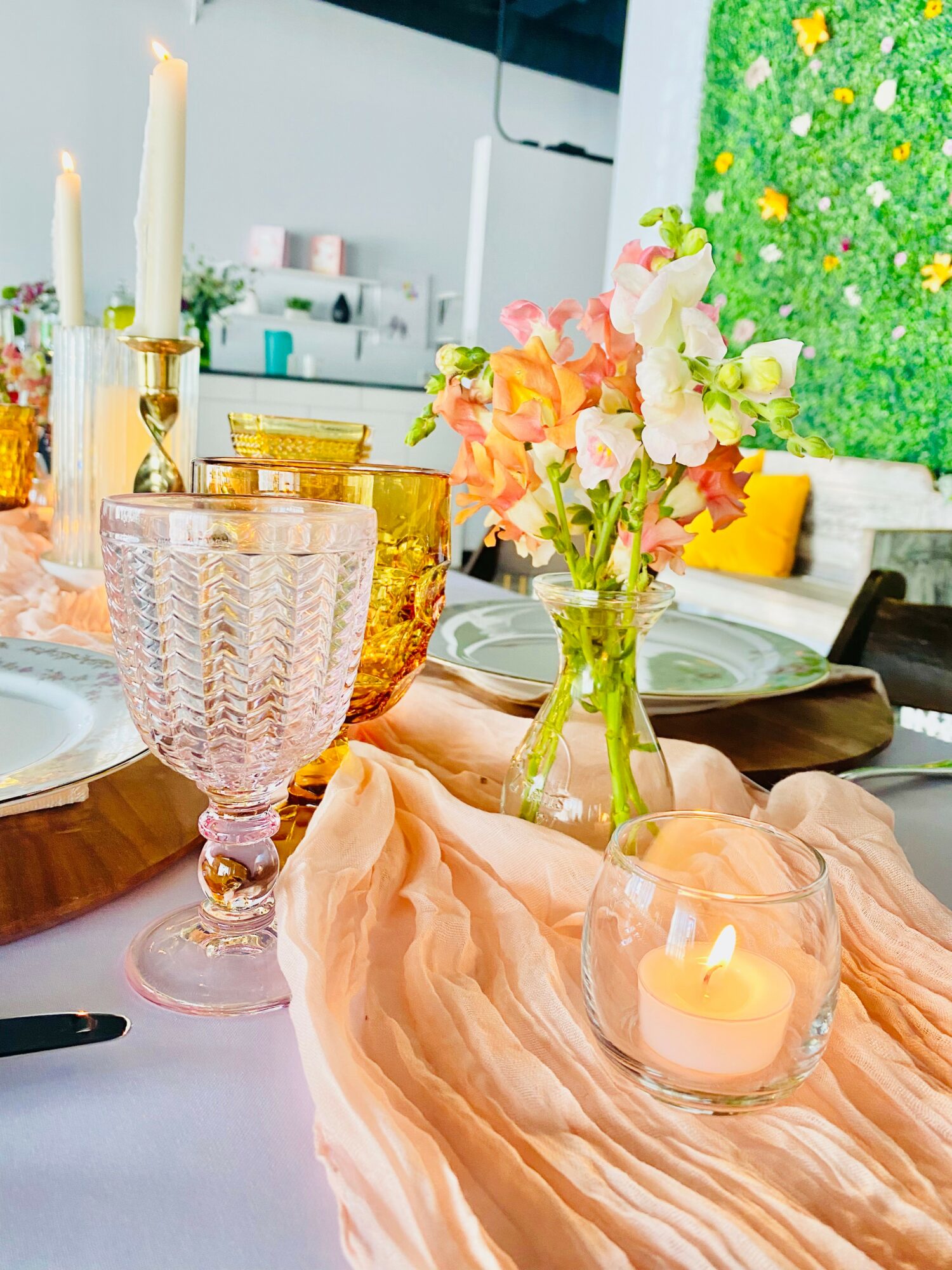 Table setting with pink cloth, glassware, candles, and floral centerpiece, with a green textured background.