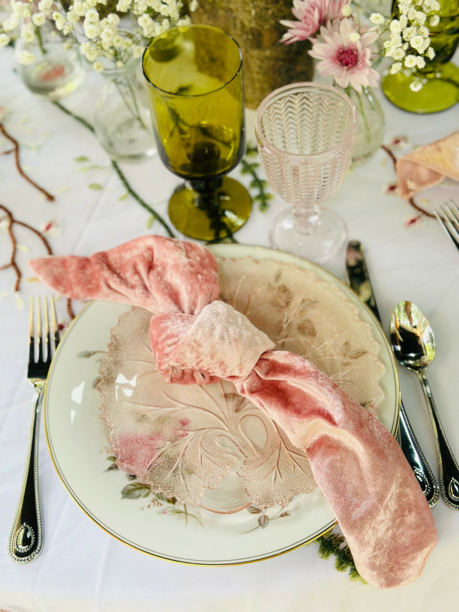 Table setting with a floral plate, pink napkin, glassware, and flowers in the background.