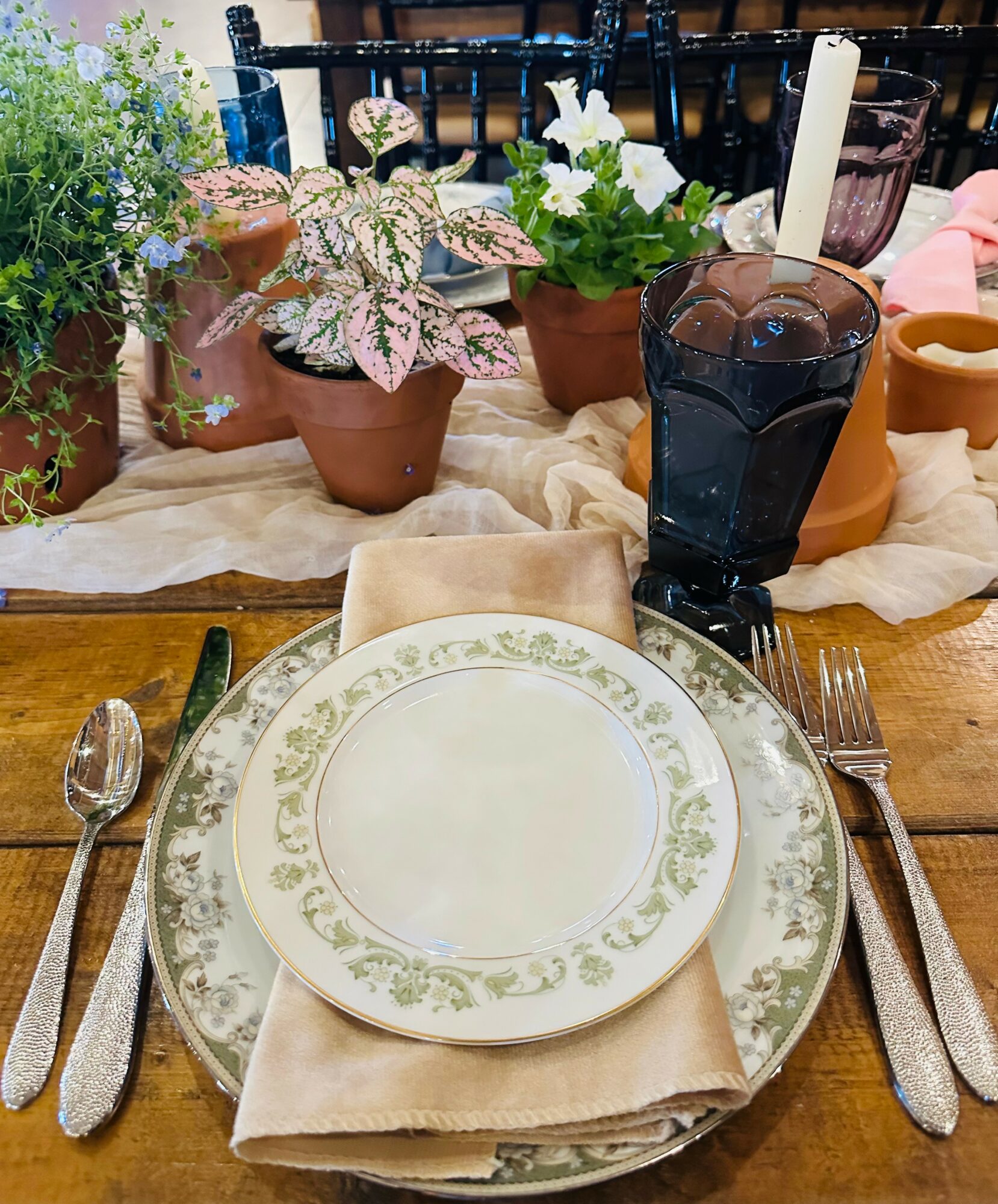 Table setting with a plate, napkin, silverware, a glass of dark beverage, and potted plants with flowers in the background.