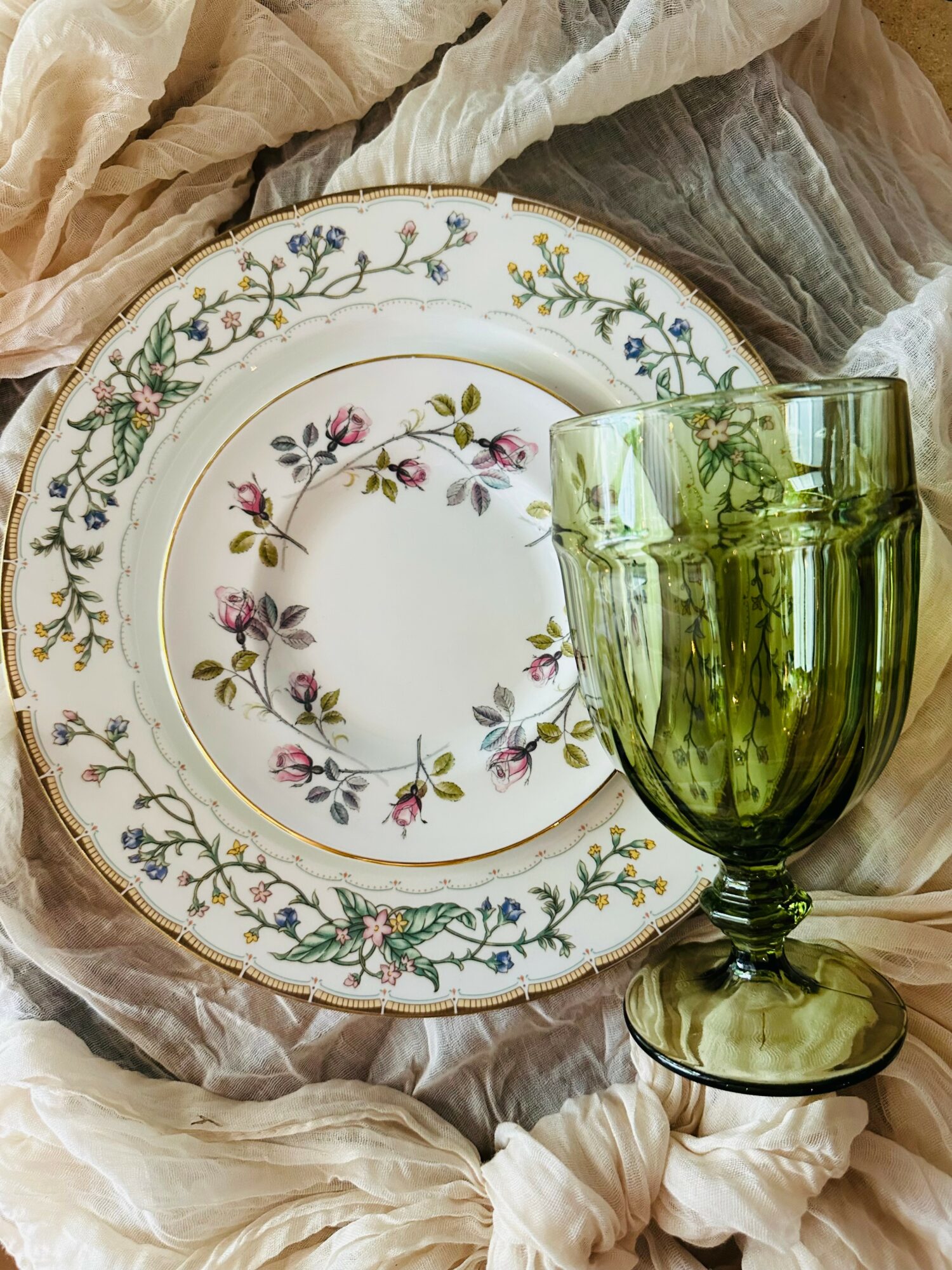 Decorative floral patterned plate with a gold rim, accompanied by a green glass goblet, on soft fabric background.
