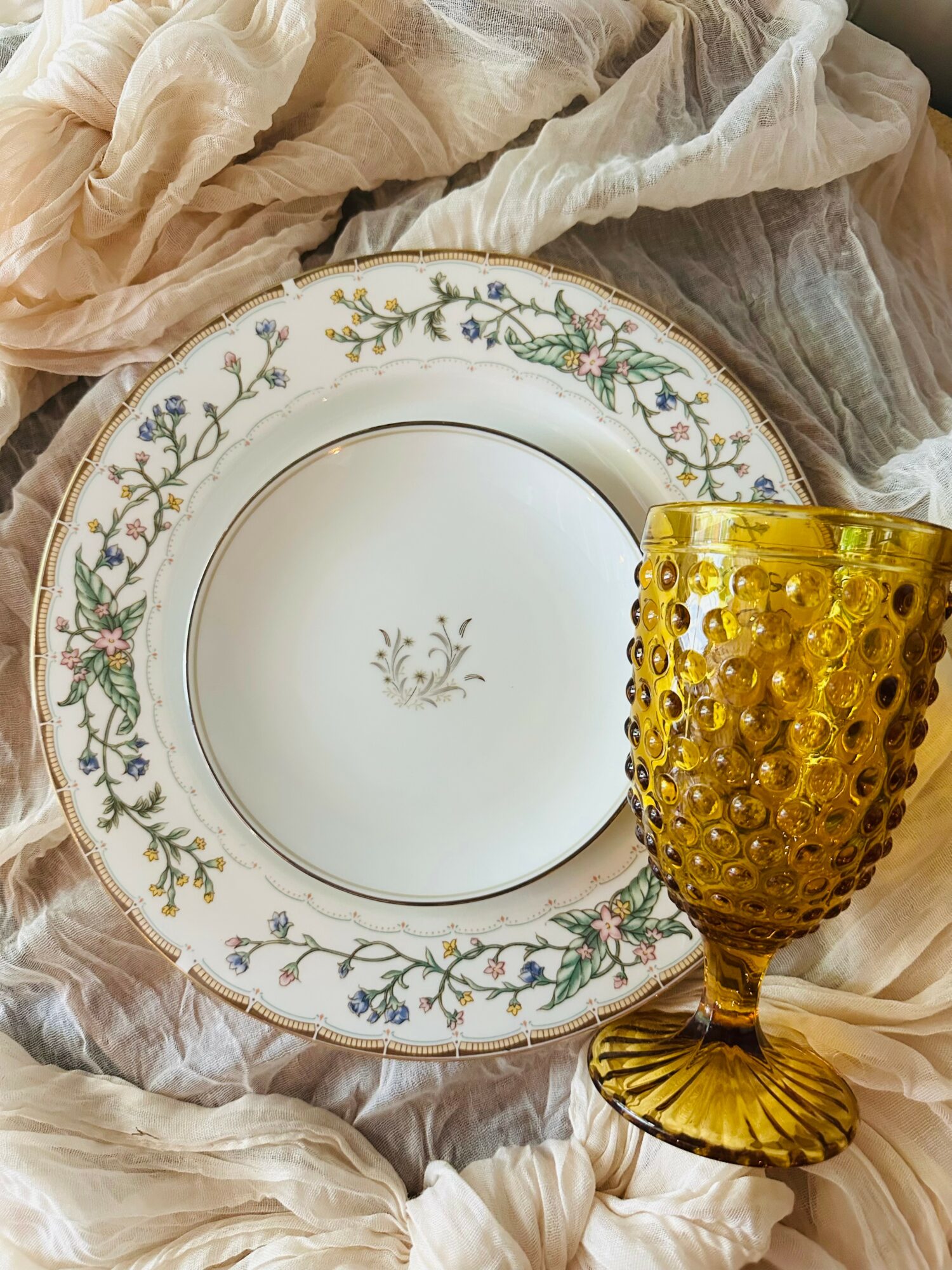 Decorative plate with floral pattern and a yellow textured glass on soft fabric background.