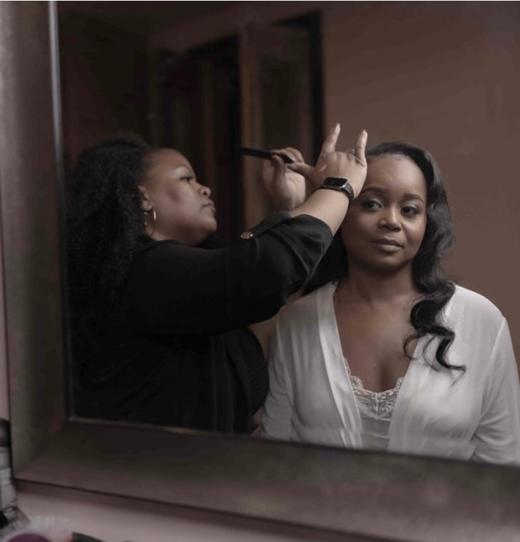 A makeup artist applies makeup to a woman reflected in a mirror, with the woman looking at the camera.