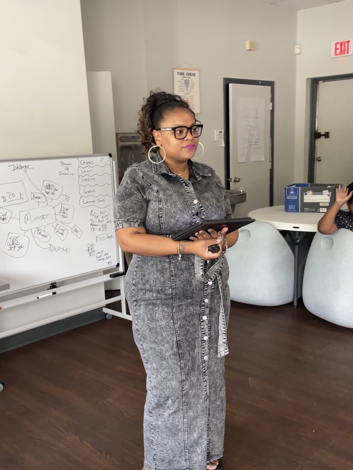 Woman with glasses and earrings holding a tablet in a room with whiteboard and chairs.