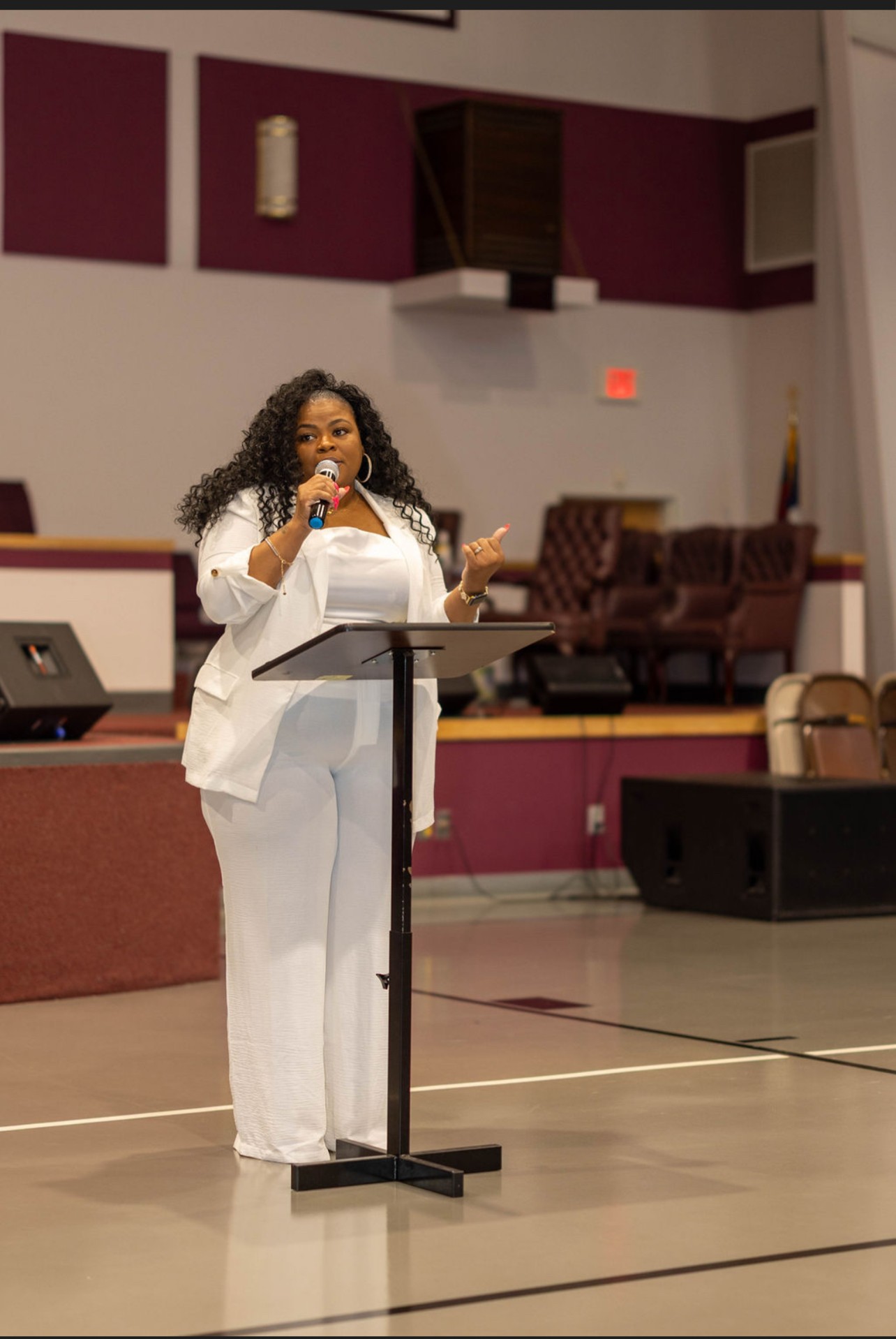 Woman speaking at a podium in a large room with chairs and stage, holding a microphone, wearing white outfit.