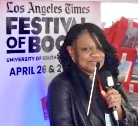 Woman speaking into microphone at festival event, background shows festival banner and red gift bags.