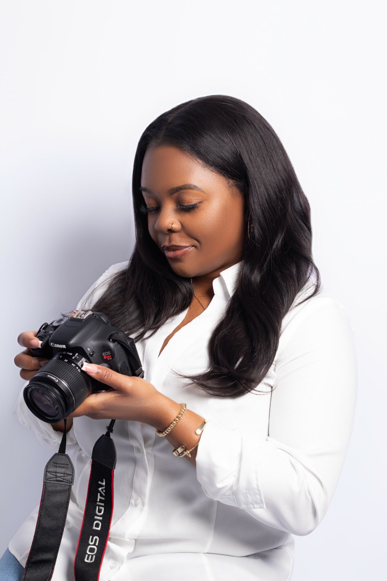 Woman with long dark hair wearing white shirt, holding a camera, looking at it, against a plain white background.
