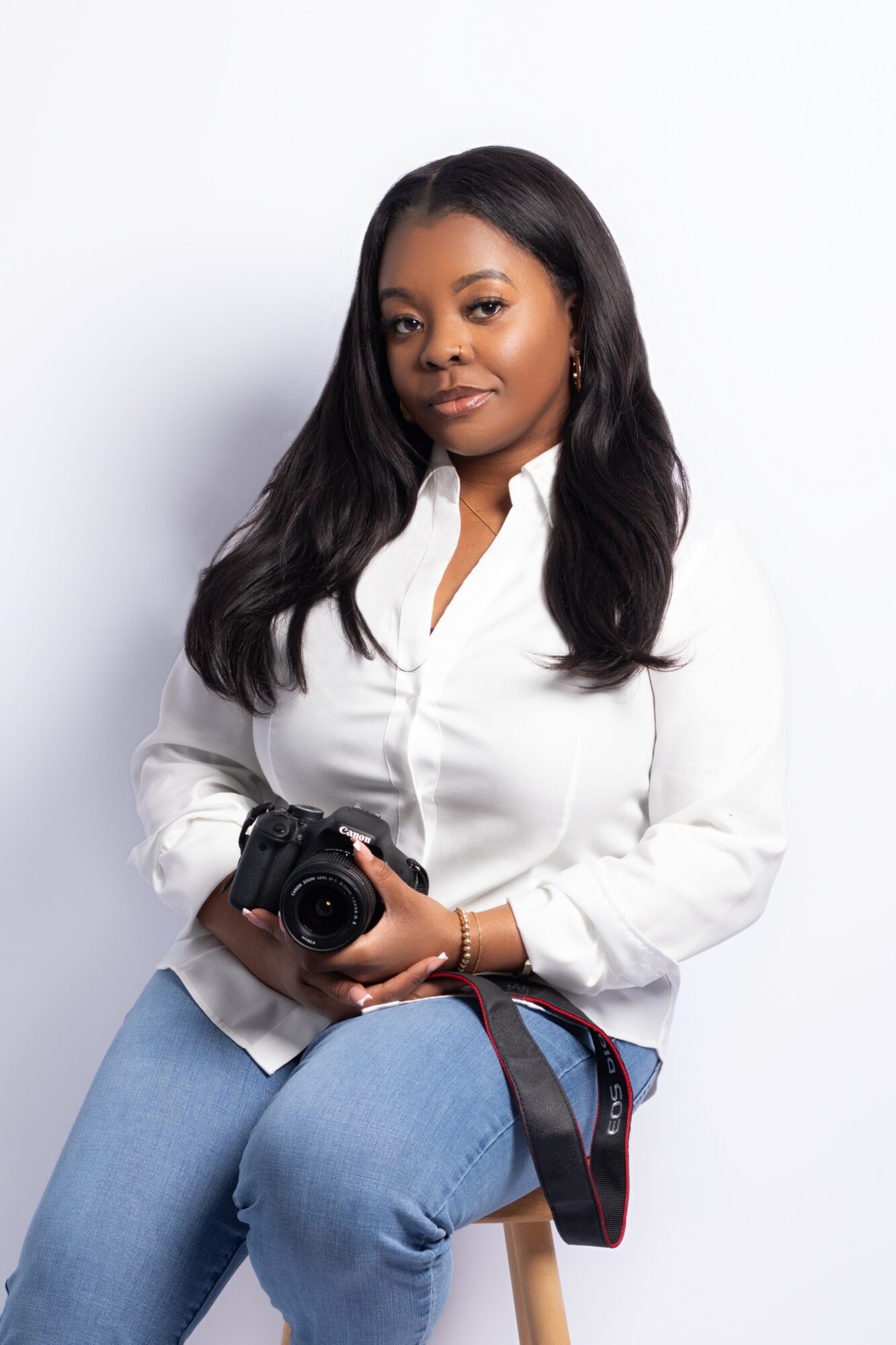 Woman with long dark hair wearing a white shirt and blue jeans, holding a camera, sitting against a white background.