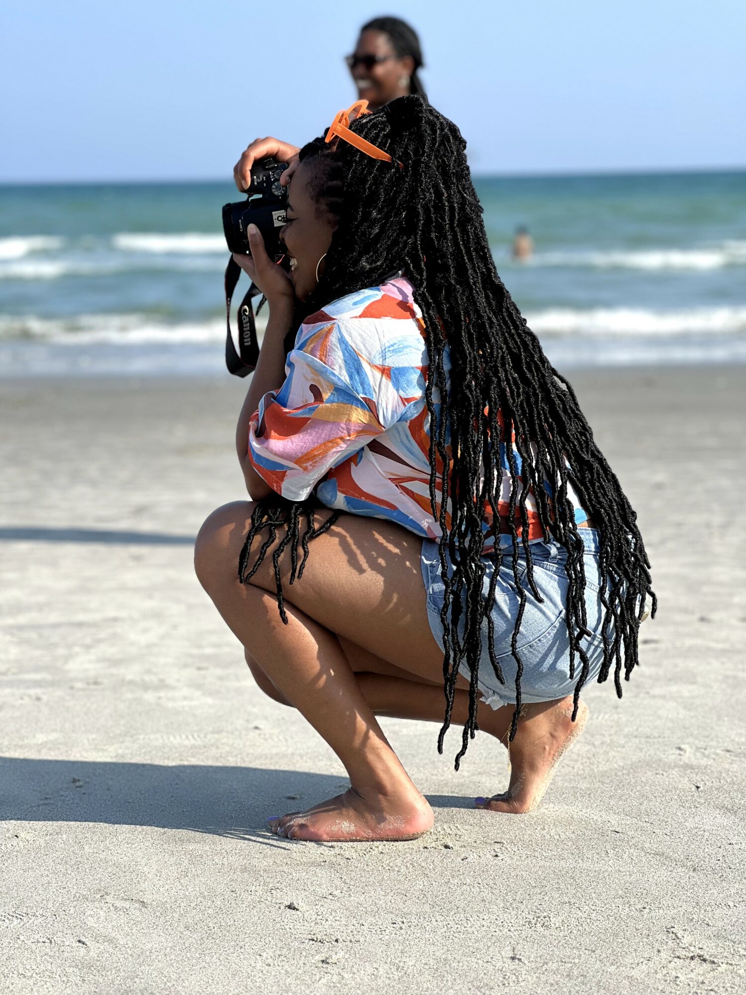 Person crouching on beach sand, holding camera, with ocean and sky in background.